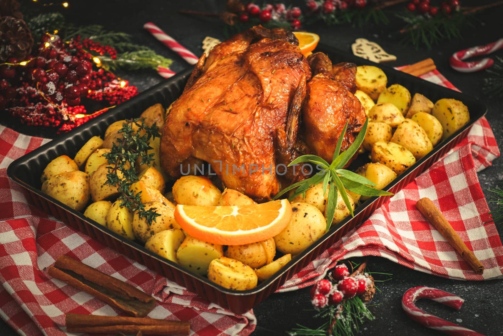 Chicken with potatoes, herbs, orange, cinnamon sticks and candy lollipops in a baking dish and christmas decoration on a black background, close-up side view.
