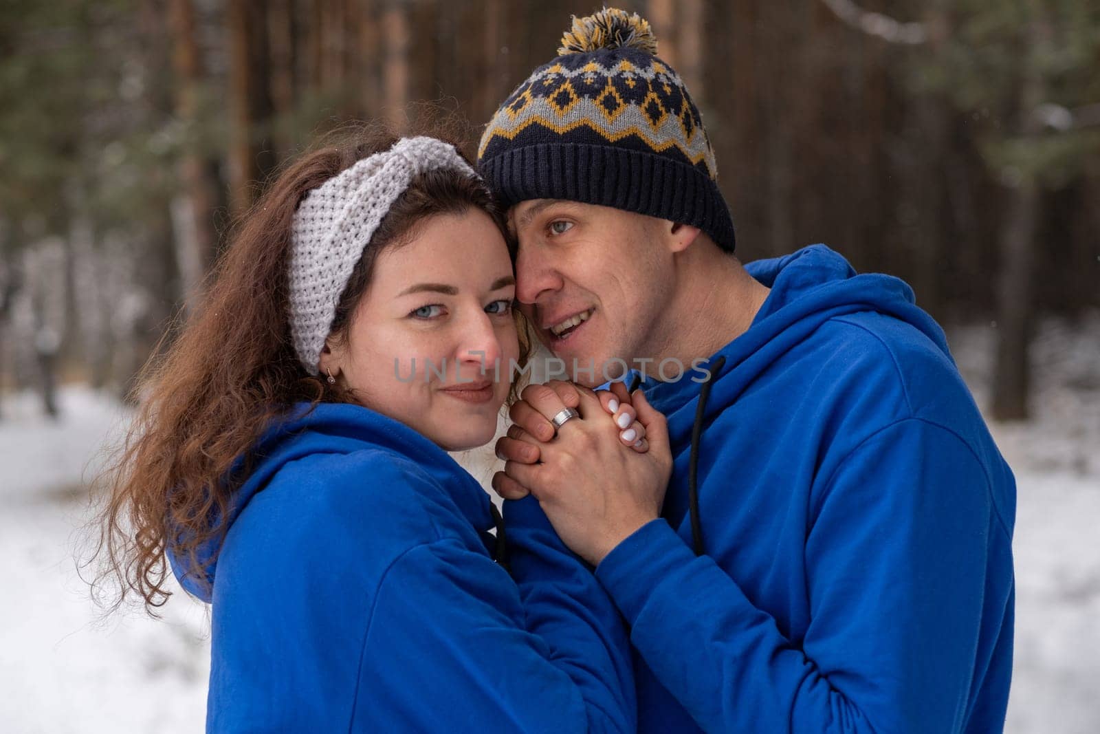 Outdoor happy couple in love posing in cold winter weather. A man and a woman in blue hoodies. Emotional young couple having fun while walking by winter forest, loving man hugging his laughing woman