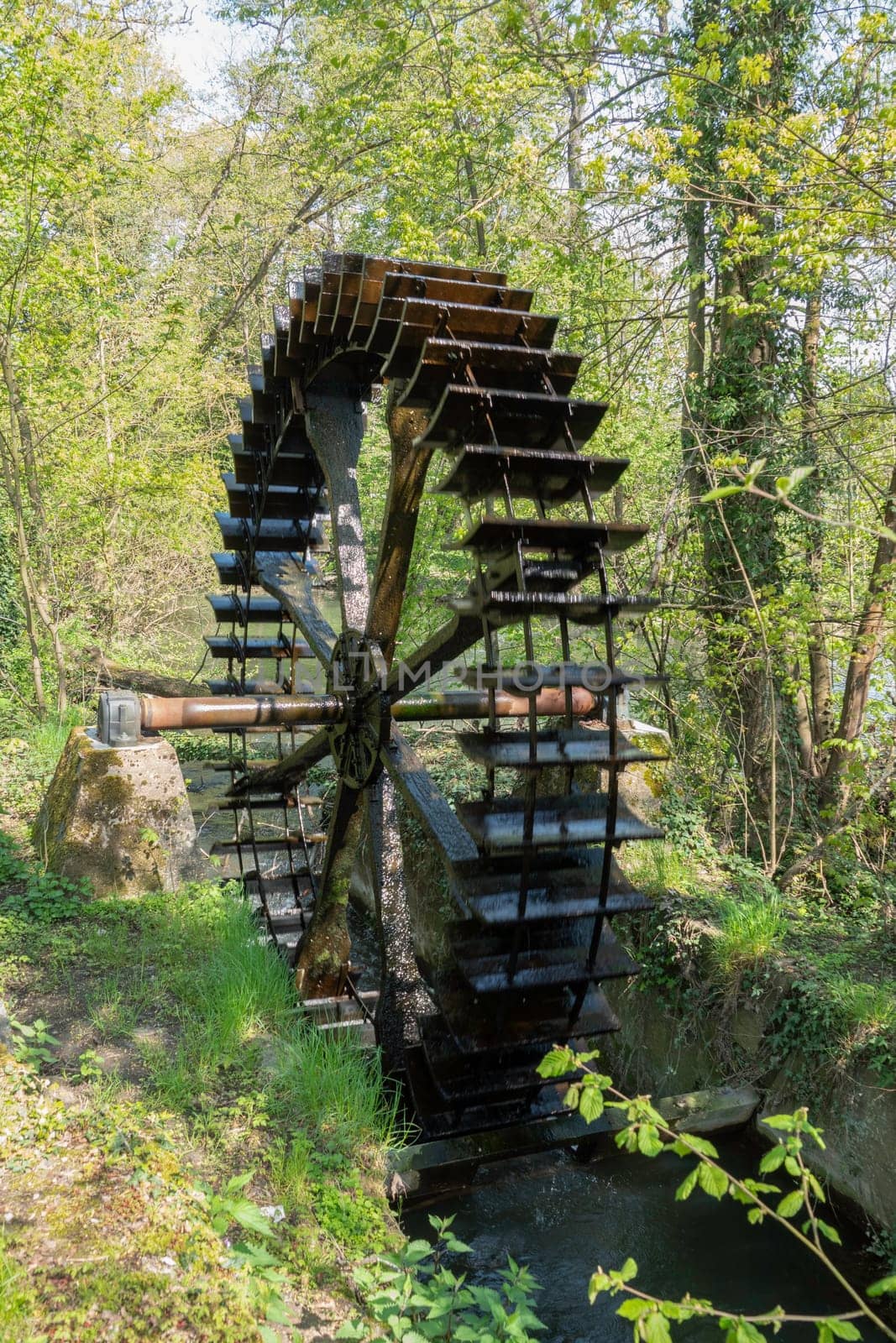 The old water wheel is located on the lake surrounded by trees and rocks on a sunny day.