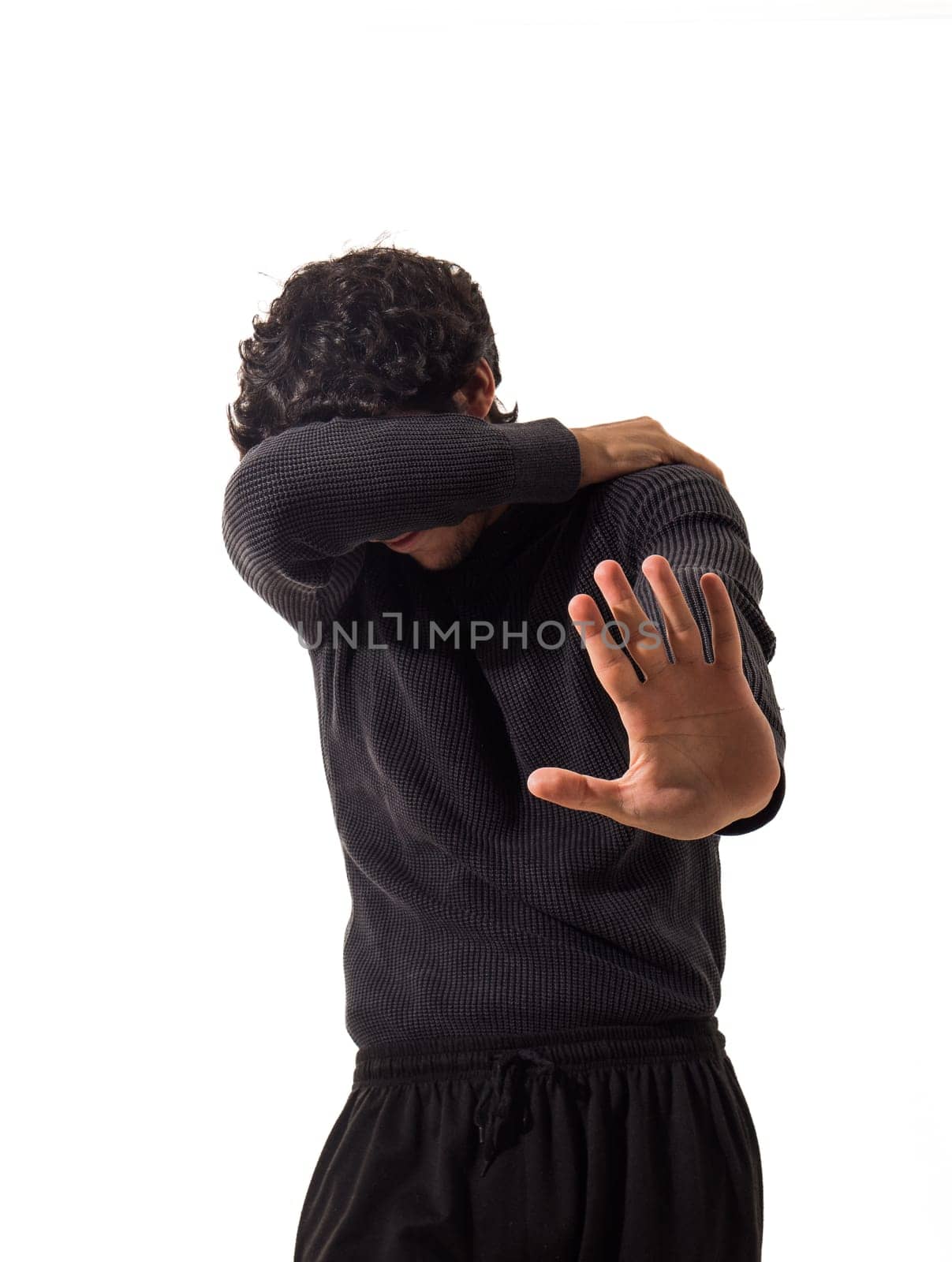 A scared man in a black sweater is protecting himself with one arm and making a stop sign with the other hand, in studio on white