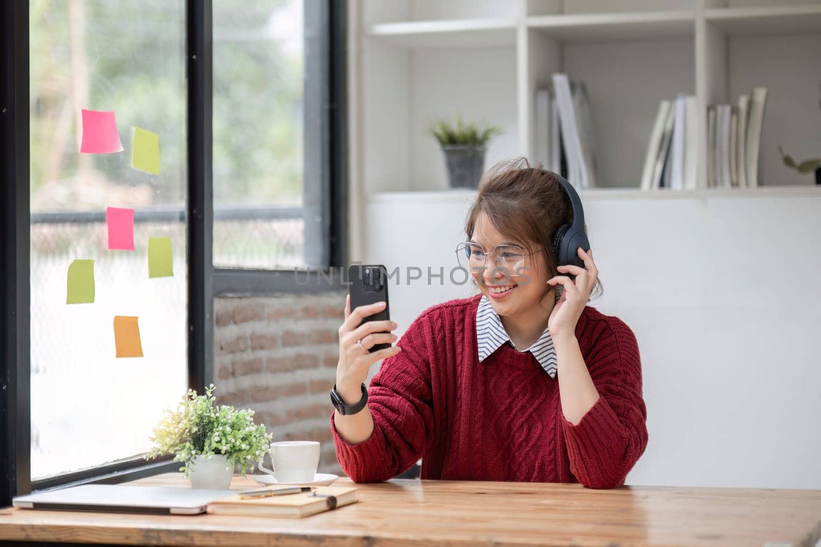 Asian female college student using laptop and phone with headphones while studying. Reading messages and greeting friends via video call.