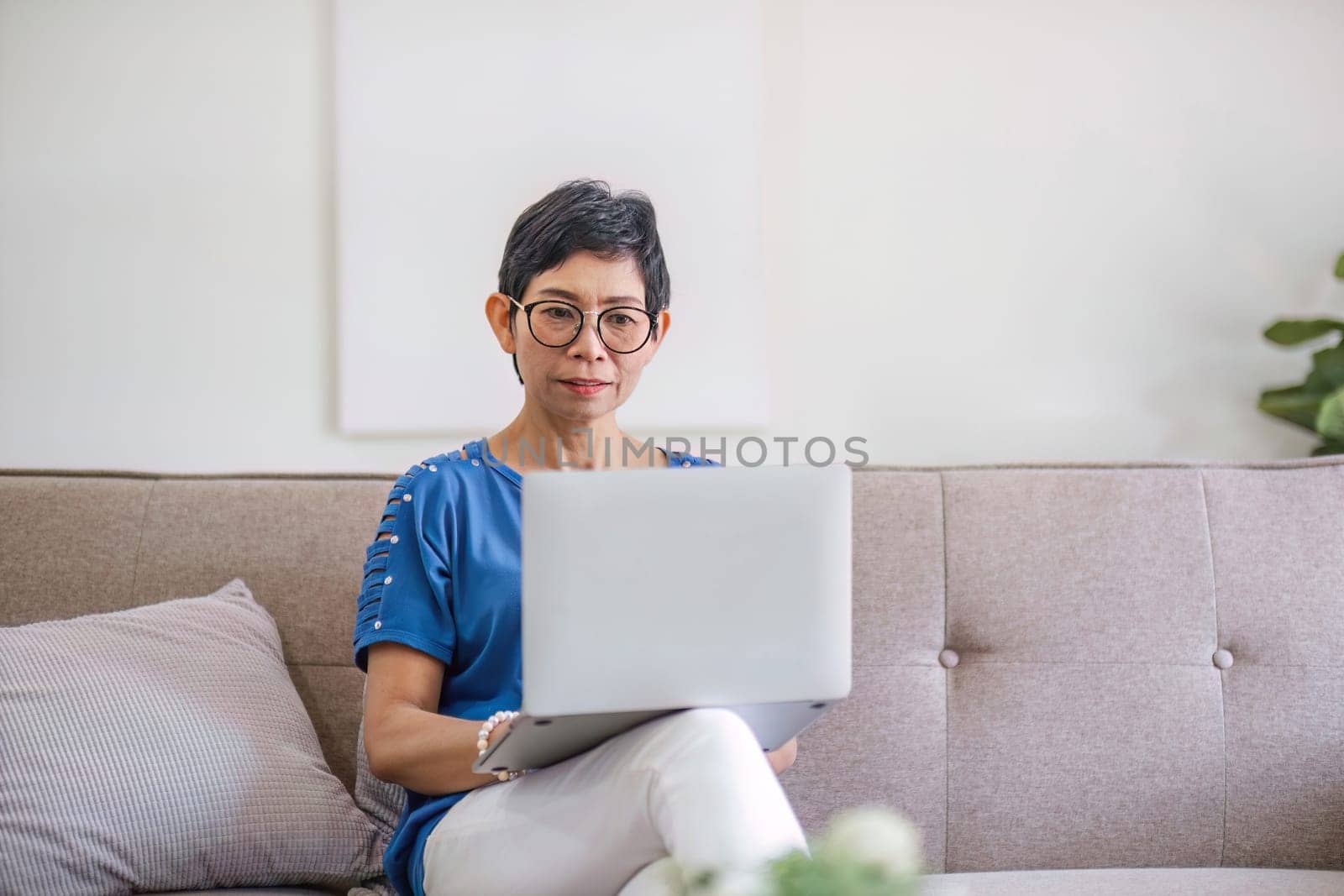 Happy Asian 60s retired woman enjoys social media on laptop while relaxing in living room..