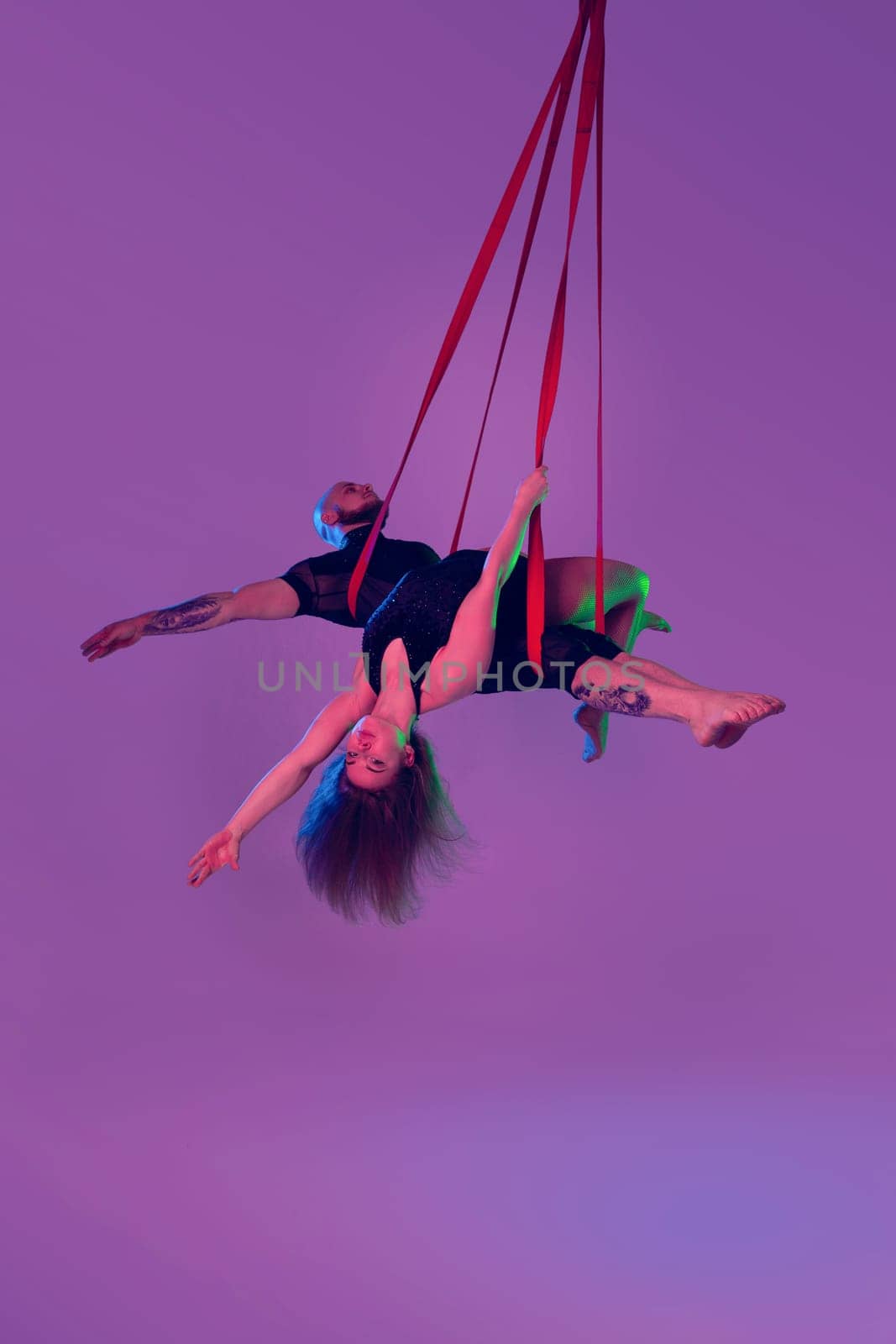 Gymnasts performance on a red canvases. Charming maiden and handsome guy in a dark sport suits are doing acrobatic elements in a studio against a colorful background. She is laying on her partner. Dancing in the air with balance.