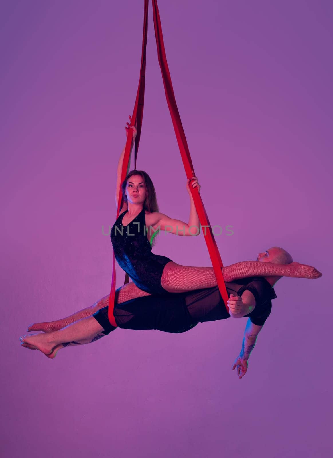 Gymnasts performance on a red canvases. Charming woman and handsome man in a dark sport suits are doing acrobatic elements showing a twine in a studio against a colorful background. Dancing in the air with balance.