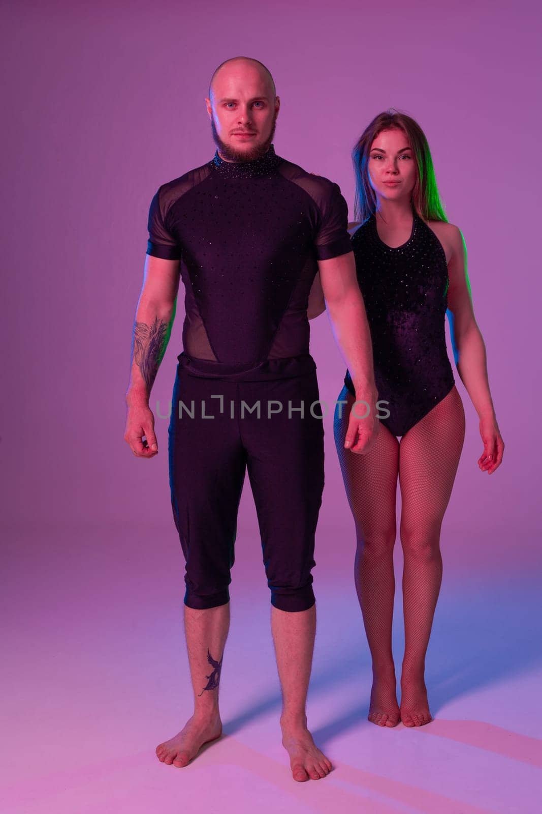 Alluring female and an athletic guy in a dark sport suits are posing in a studio against a colorful background. Gymnasts performance.