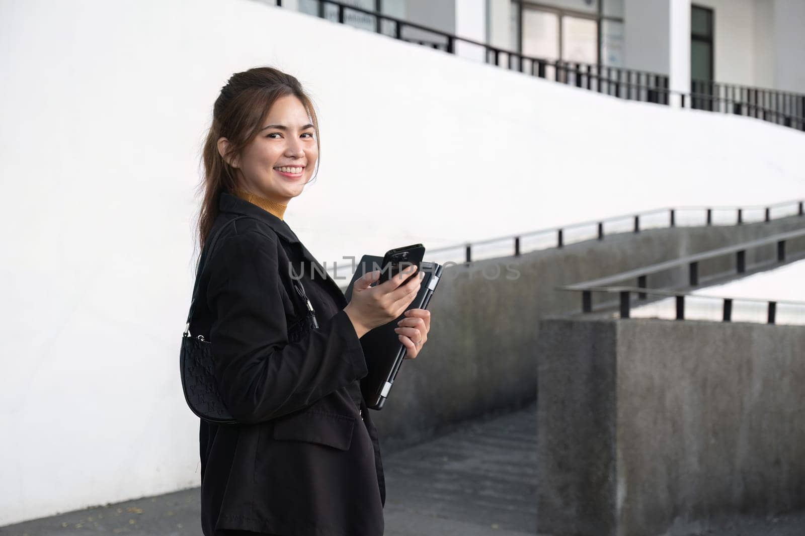 Attractive smiling Asian businesswoman wearing a suit standing in the city using an application on her mobile phone Read news on your smartphone fast connection Check out the outdoor mobile app by wichayada
