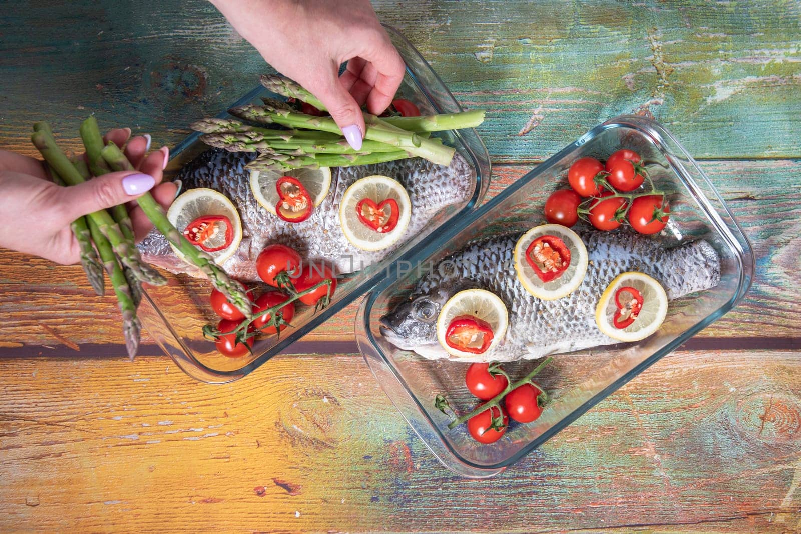 woman's hand puts green asparagus on fresh large dorado fish, tilapia, carp in a baking dish with cherry tomatoes,step by step recipe instructions, top view on an old wooden table, High quality photo