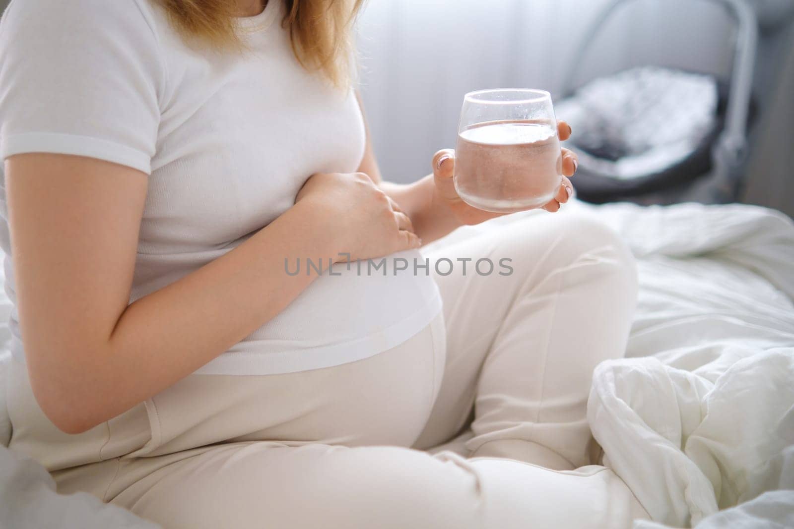 Pregnant woman sits on the bed, tenderly embracing her growing belly and holding glass of water.