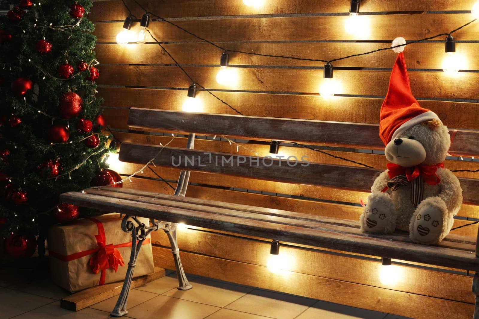 A sad teddy bear in a Santa hat sits on a bench next to a New Year tree, a gift box, against the backdrop of festive glowing lamps.