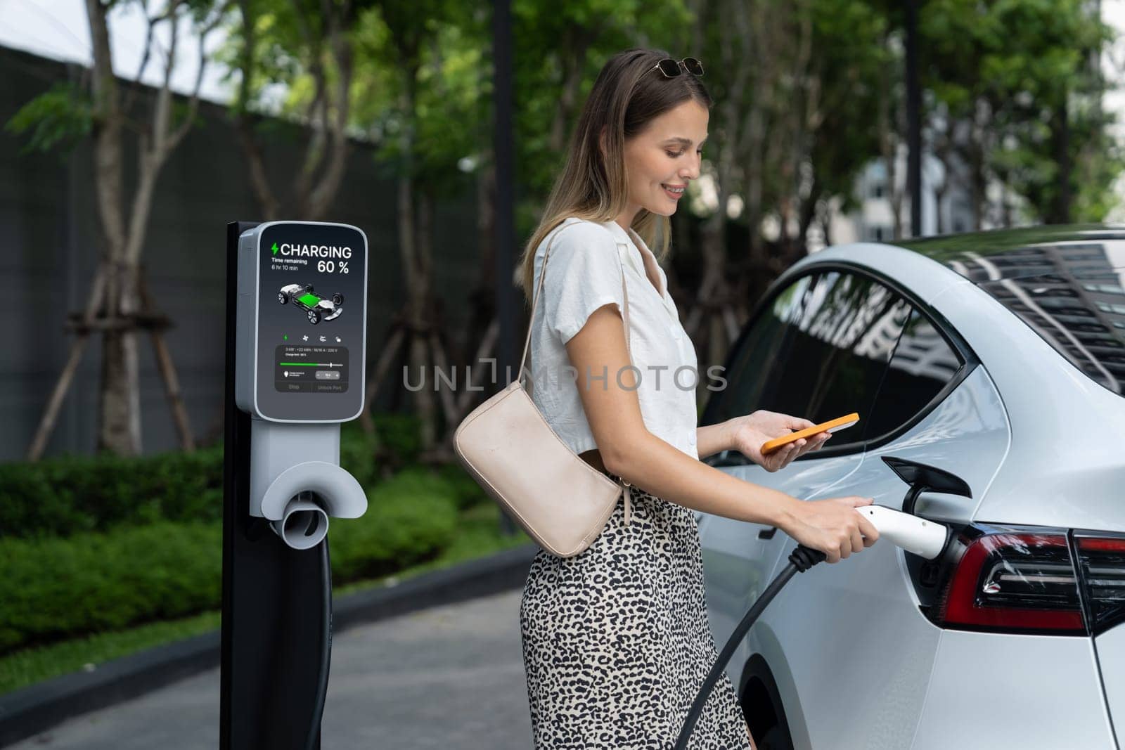 Young woman travel with EV electric car charging in green sustainable city outdoor garden in summer. Urban sustainability lifestyle by green clean rechargeable energy of electric BEV vehicle innards