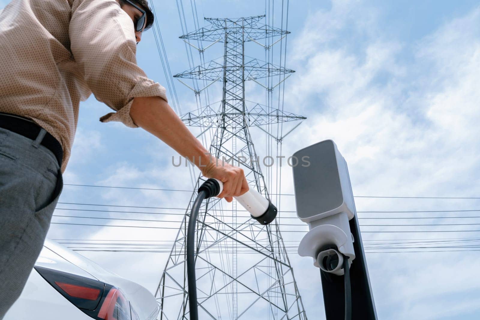 Man recharge EV electric car battery at charging station connected to electrical power grid tower on sky background as electrical industry for eco friendly vehicle utilization. Expedient