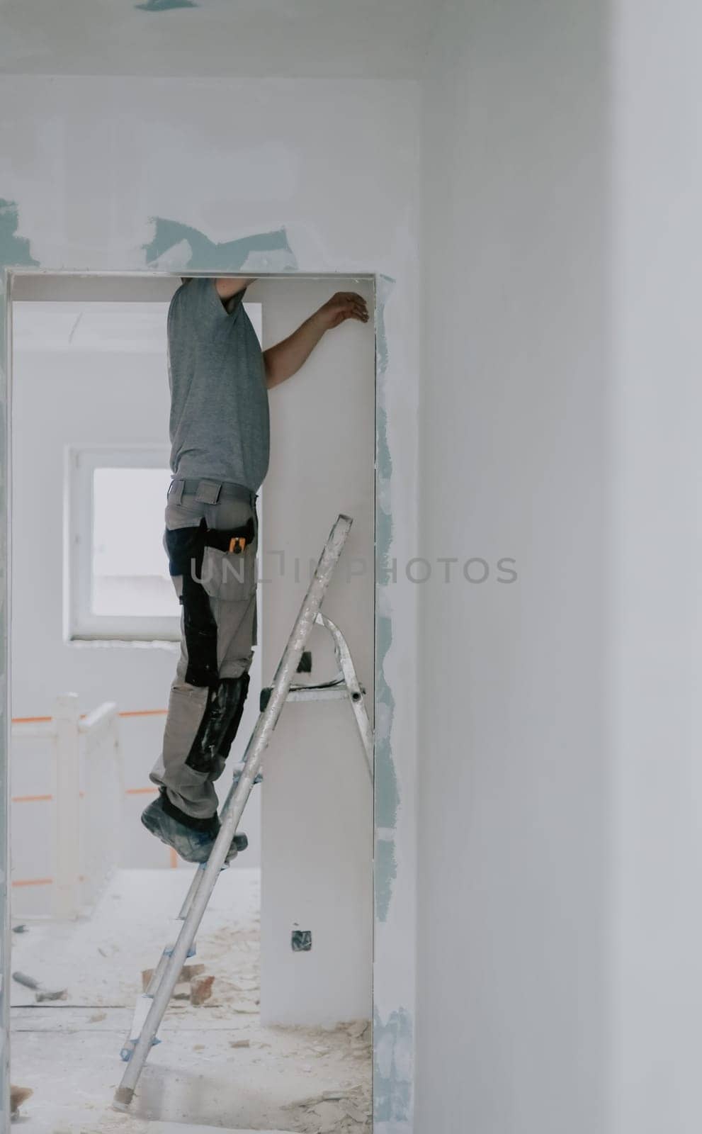 One young Caucasian man in a gray T-shirt clears old putty from the top of a doorway with one hand using an ax while standing sideways on a stepladder, close-up side view with selective focus. Construction work concept.