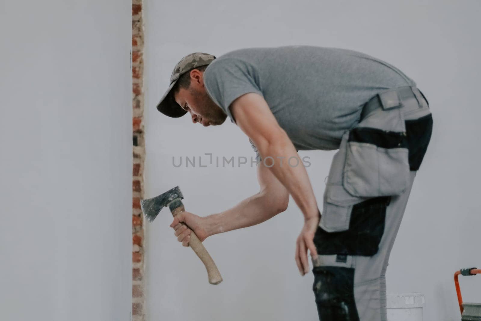 One young Caucasian man in a gray T-shirt and cap is cleaning old putty from the side of a doorway with an ax while standing at an angle, close-up side view with selective focus. Construction work concept.