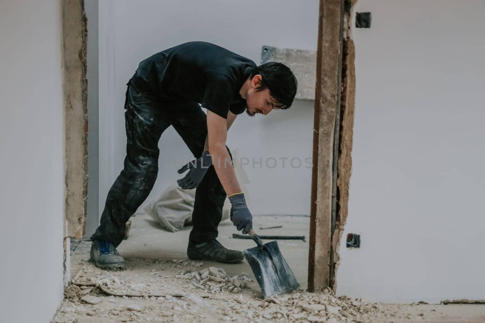 One young handsome Caucasian man, standing bent over, collects construction waste from the floor using a shovel, side view close-up.