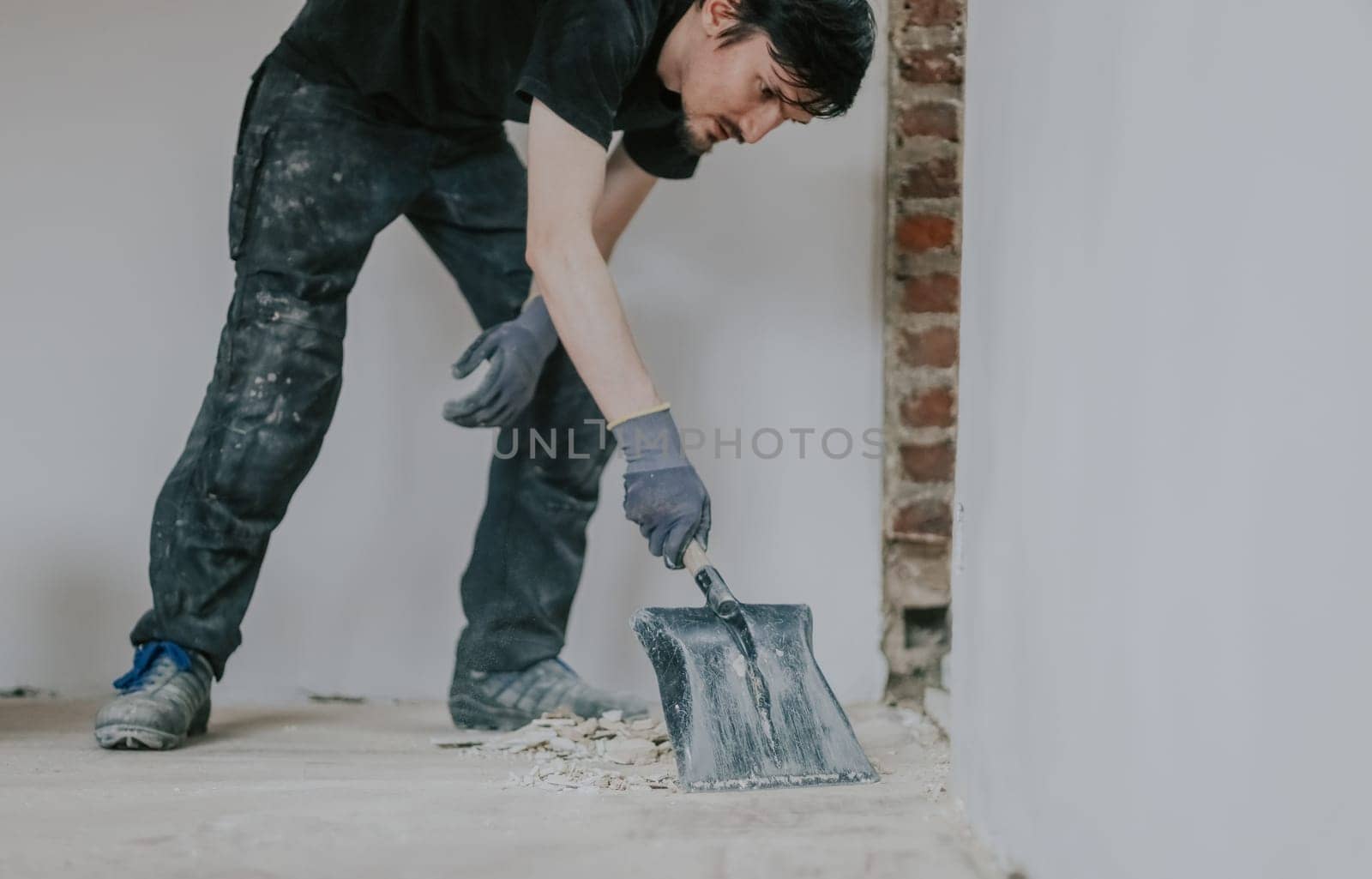 One young handsome brunette Caucasian woman in a black uniform stands, bending over, collecting construction debris from the floor with a shovel, close-up side view.