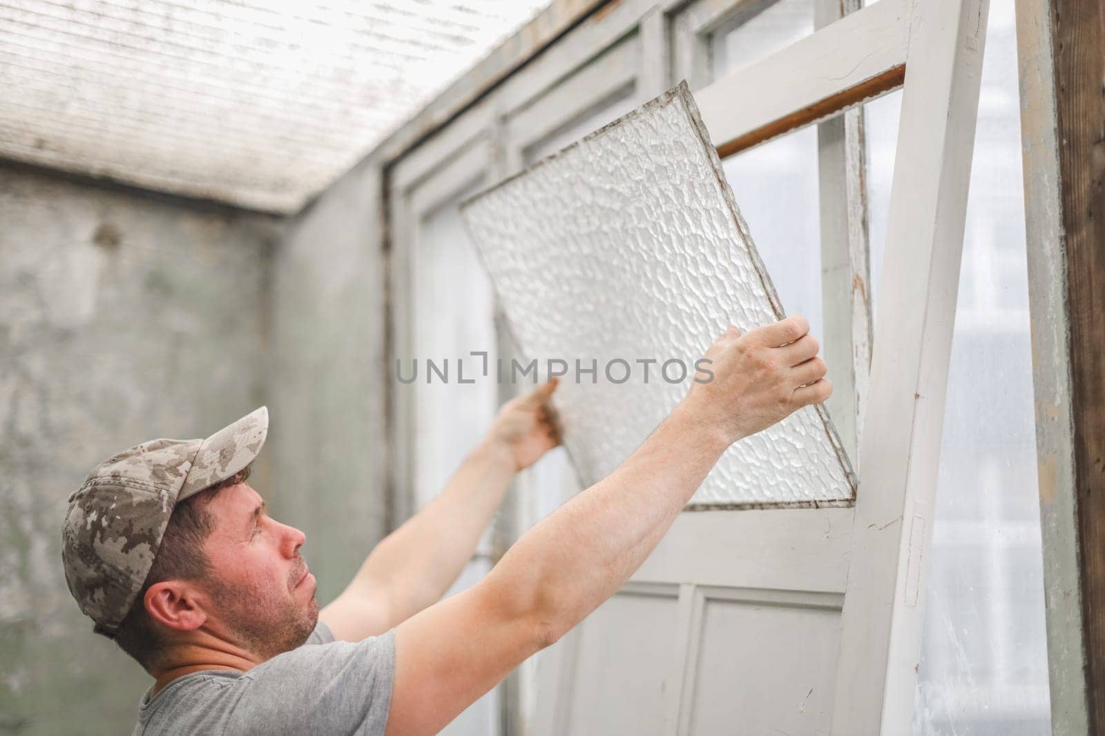 One young handsome Caucasian man in a gray T-shirt and cap removes with both hands the corrugated glass from an old door while standing in a room where renovations are underway, close-up side view.