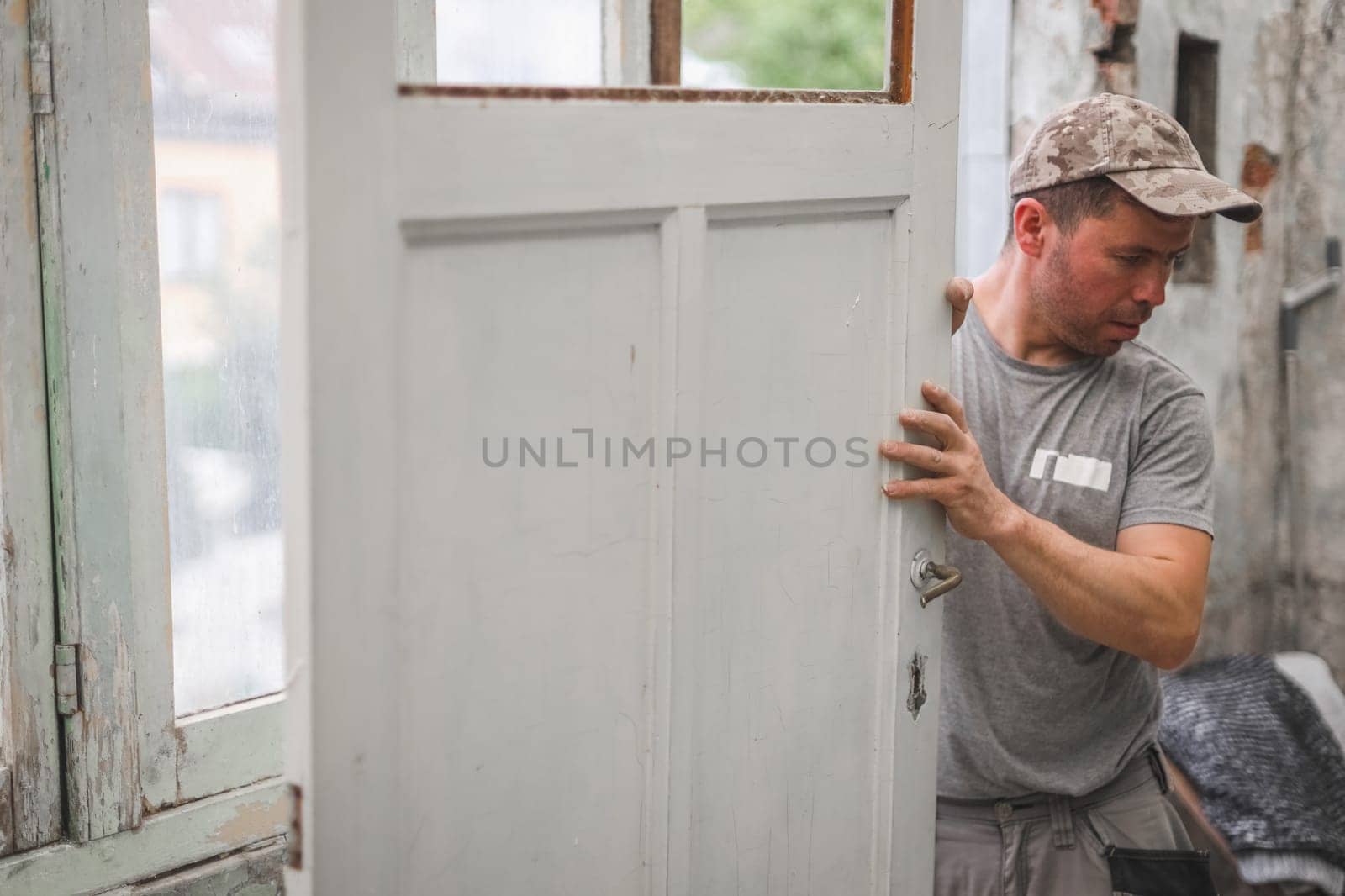 One young handsome Caucasian man in a gray T-shirt and cap holds with both hands an old wooden door while standing in a room where renovations are underway, close-up side view.