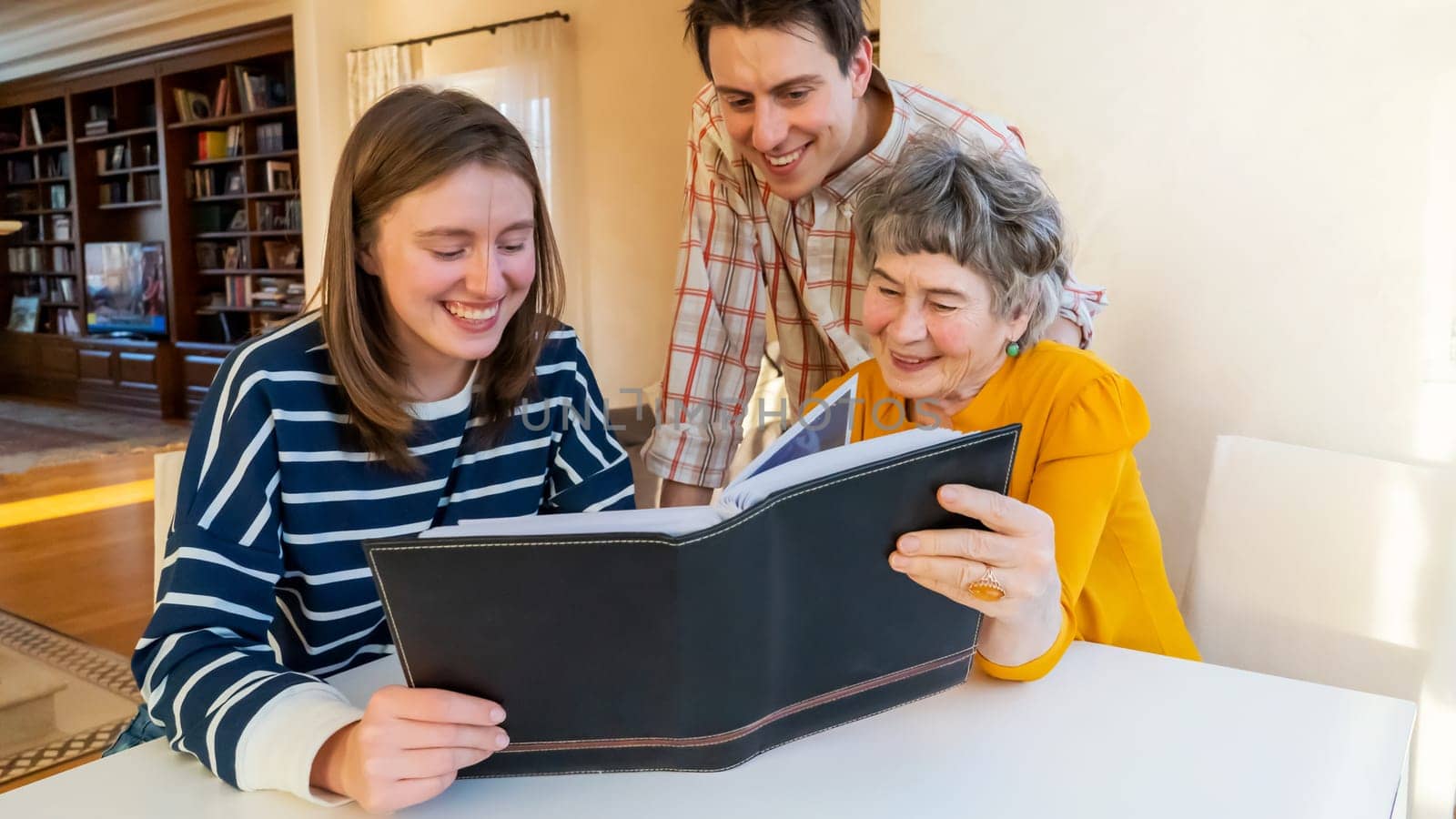 Family smiles happily and spends time together at home, young girl and a man look at photo album with their grandmother, help an elderly woman remember her youth, visit and take care of grandparents.