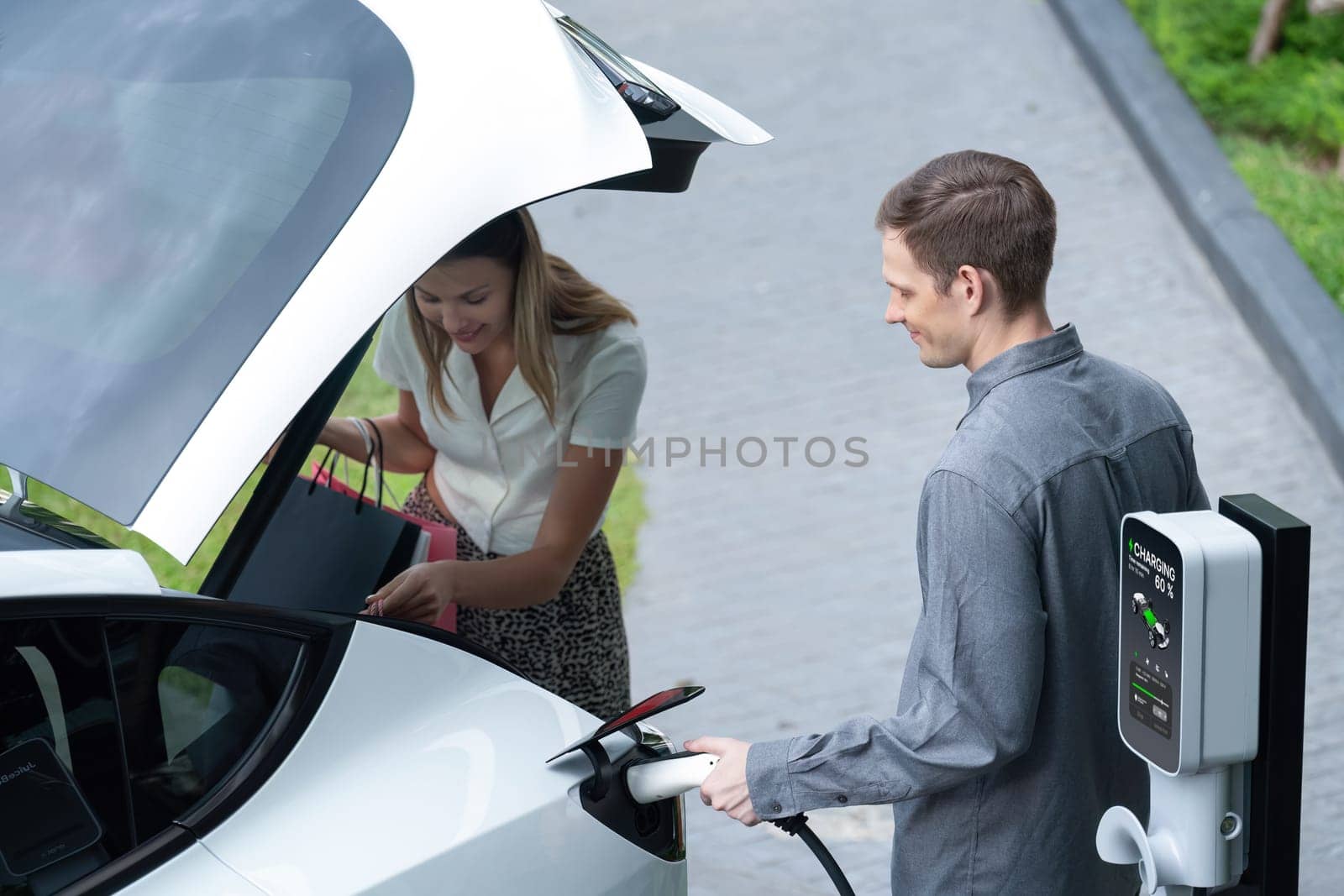 Young couple travel with EV electric car charging in green sustainable city outdoor garden in summer shows urban sustainability lifestyle by green clean rechargeable energy of electric vehicle innards