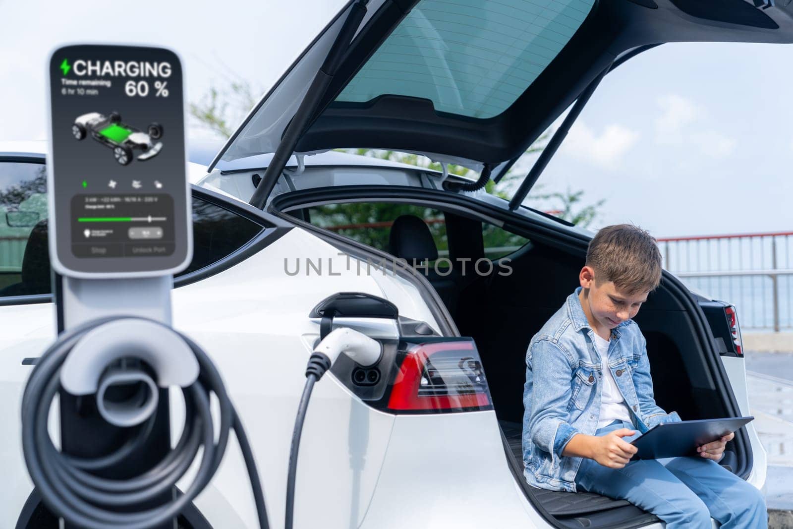 Little boy sitting on car trunk, using tablet while recharging eco-friendly car from EV charging station. EV car road trip travel as alternative vehicle using sustainable energy concept. Perpetual