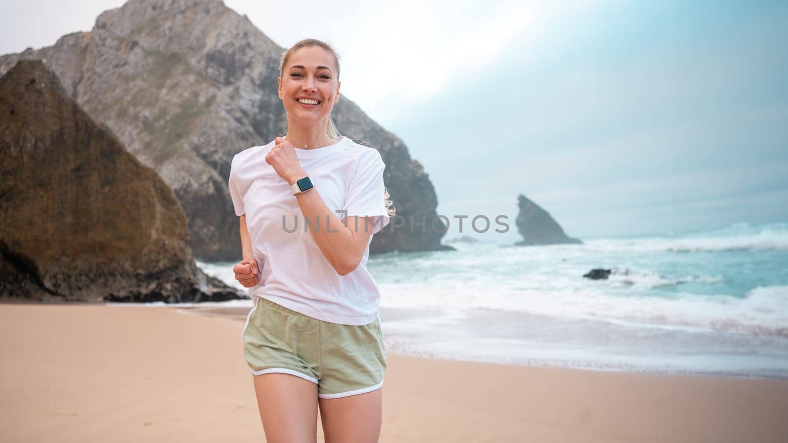Woman smiling and jogging on beach with sky and rocks in background. Woman in white t-shirt and light green athletic shorts jog on beach. Athletic smiling woman running on sandy beach at seaside