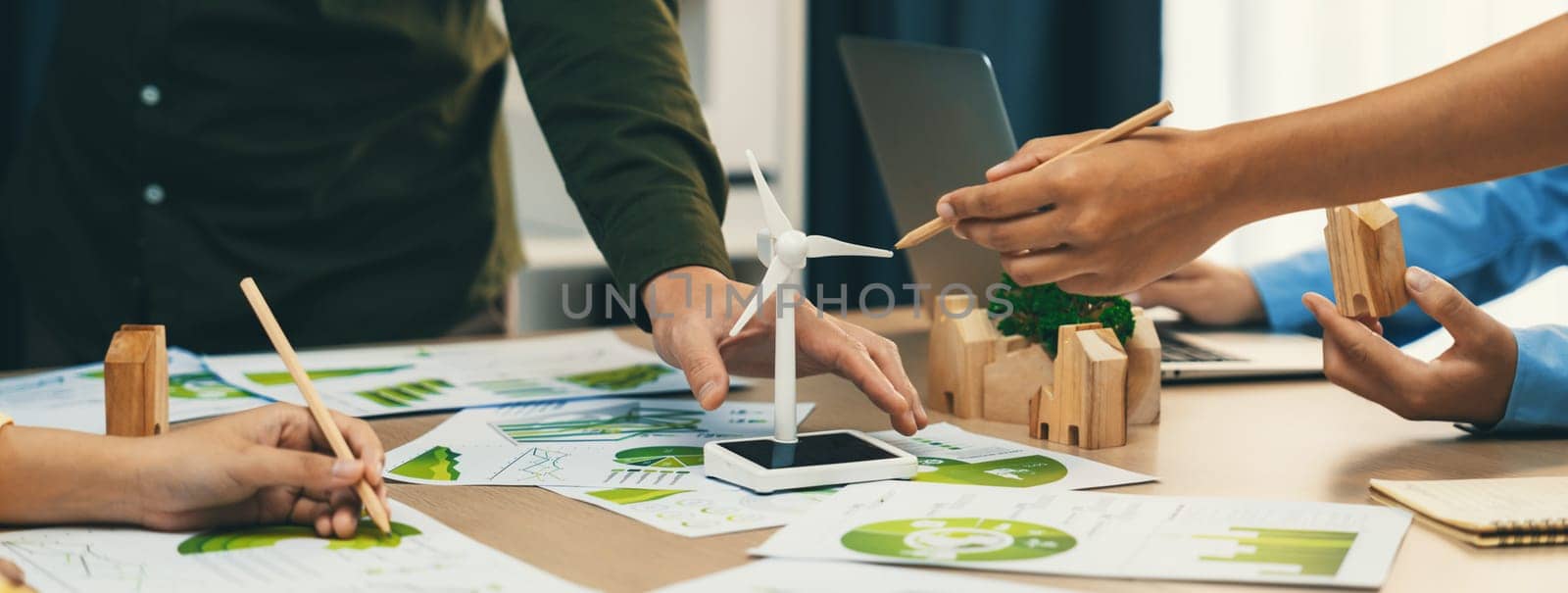 Windmill model represented renewable energy and wooden block represented eco city was placed on green business meeting table with environmental document scatter around. Front view. Delineation.