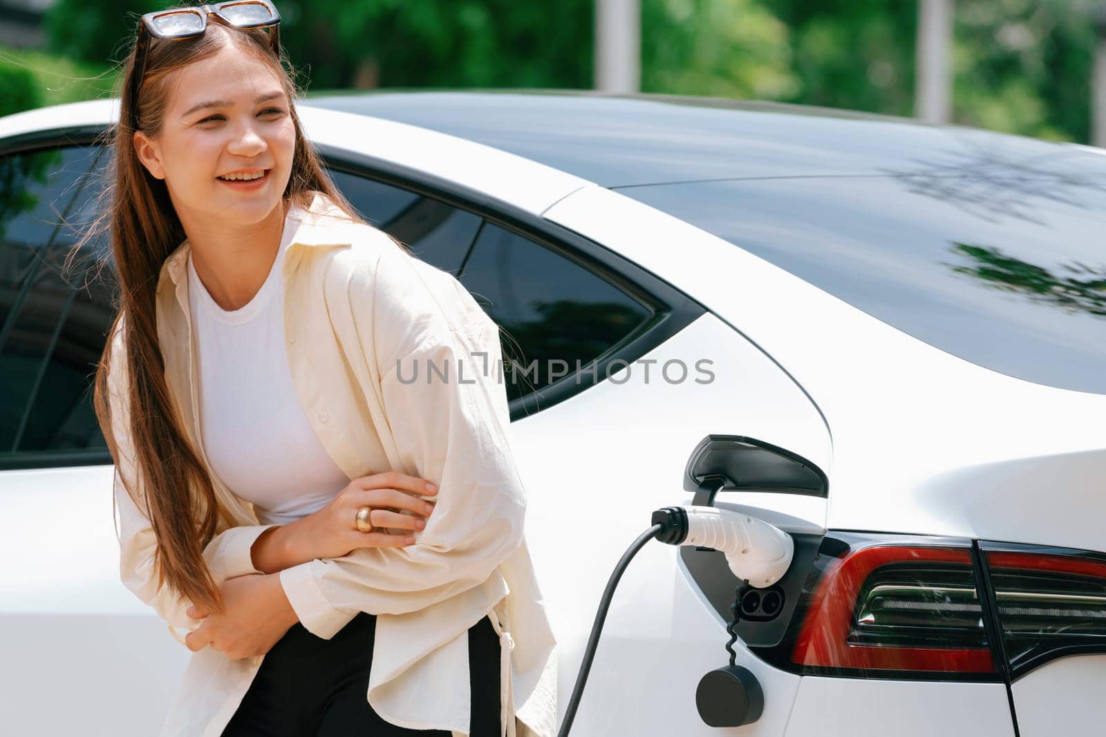 Young woman recharge her EV electric vehicle at green city park. Expedient by biancoblue