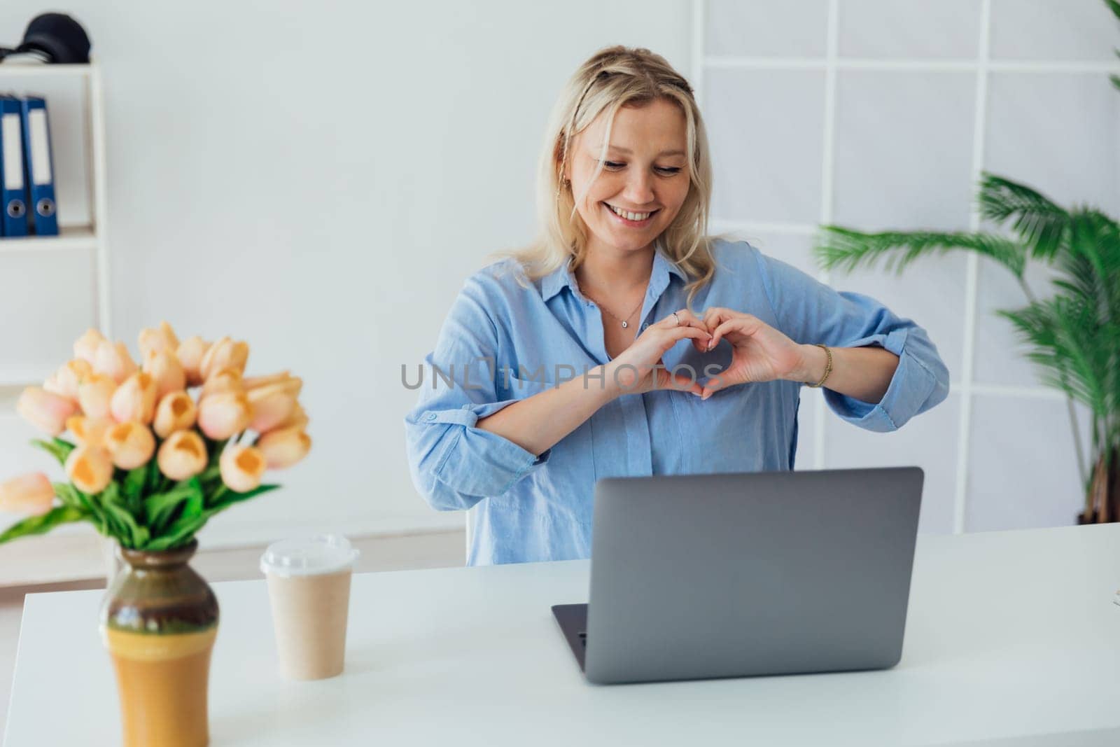 Friendly professional woman making a heart shaped sign indicating positivity and love. Corporate female employee sitting at office desk showing positive feelings while working.