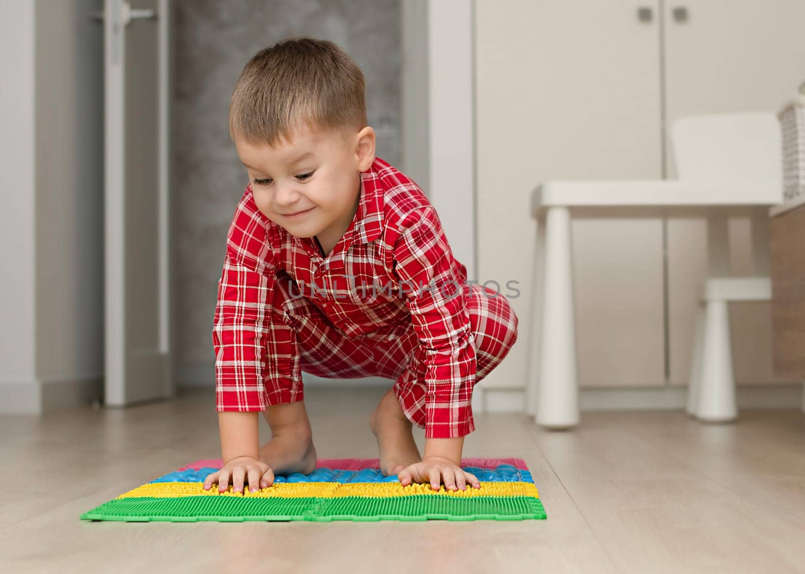 Sport and health concept. A little boy 4 years old in red checkered pajamas is working out on a multi-colored massage orthopedic mat with spikes in a home interior. Close-up. by ketlit