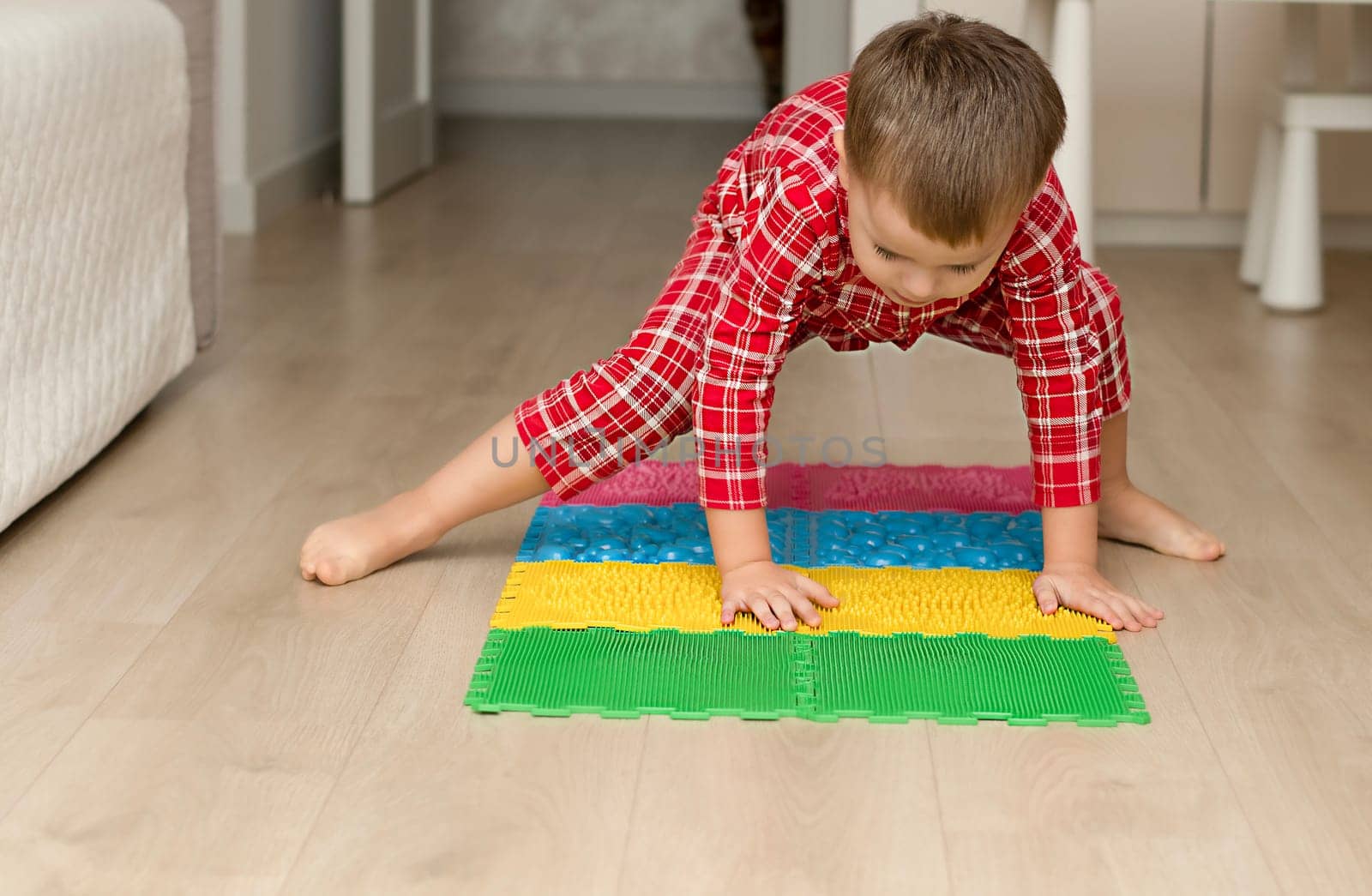 Sport and health concept. A little boy 4 years old in red checkered pajamas is having fun on a multi-colored massage orthopedic mat with spikes in a home interior. Close-up. Soft focus.