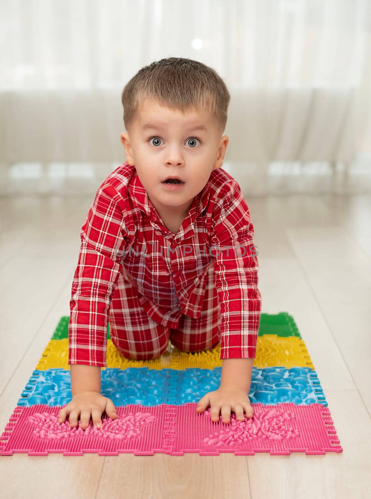 Sport and health concept. A little boy 4 years old in red checkered pajamas is working out on a multi-colored massage orthopedic mat with spikes in a home interior. Close-up. by ketlit