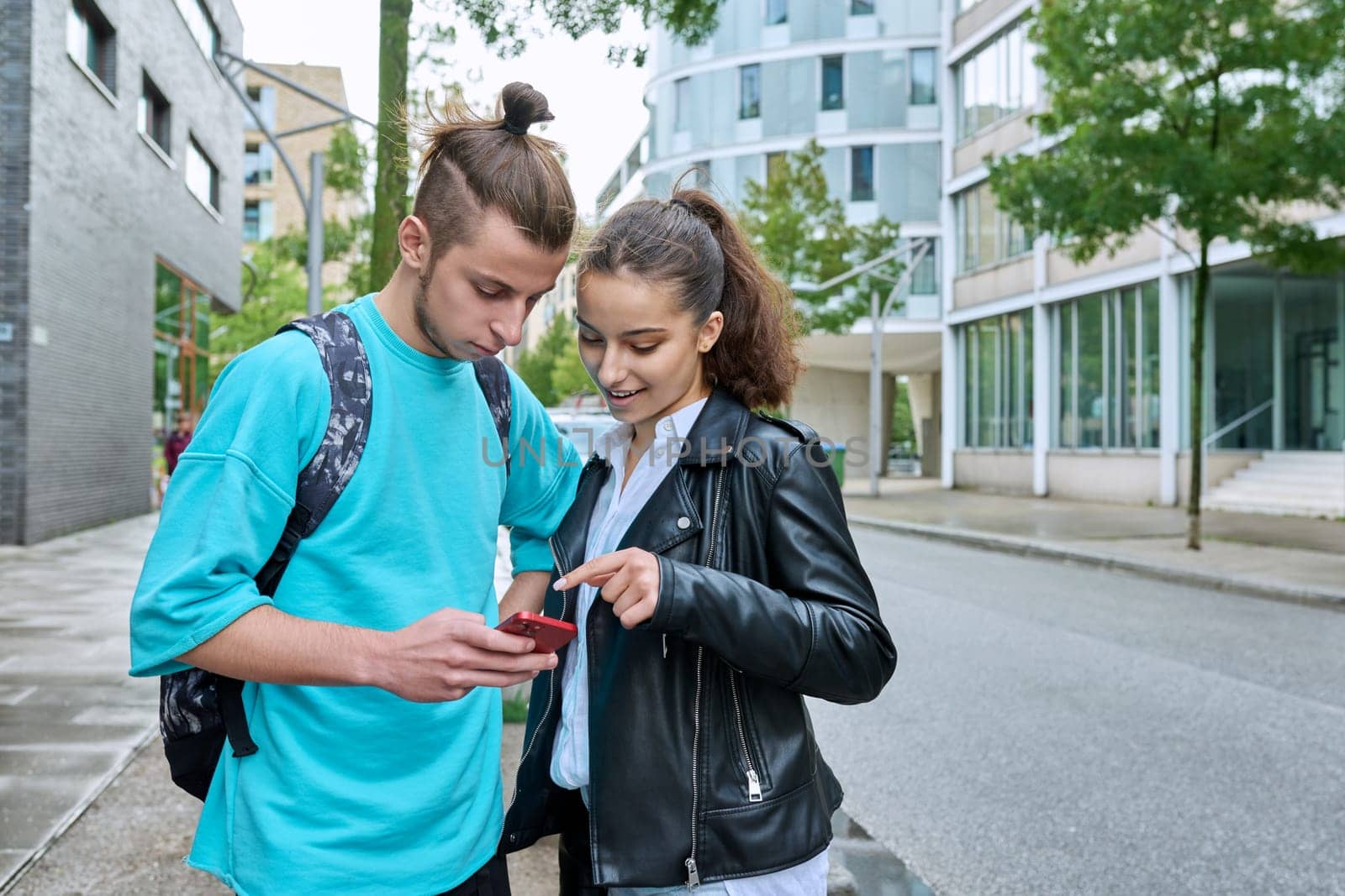 Teen friends guy and girl standing together holding smartphones looking at screen using mobile phones outdoor on city. Internet digital technology applications for leisure study communication