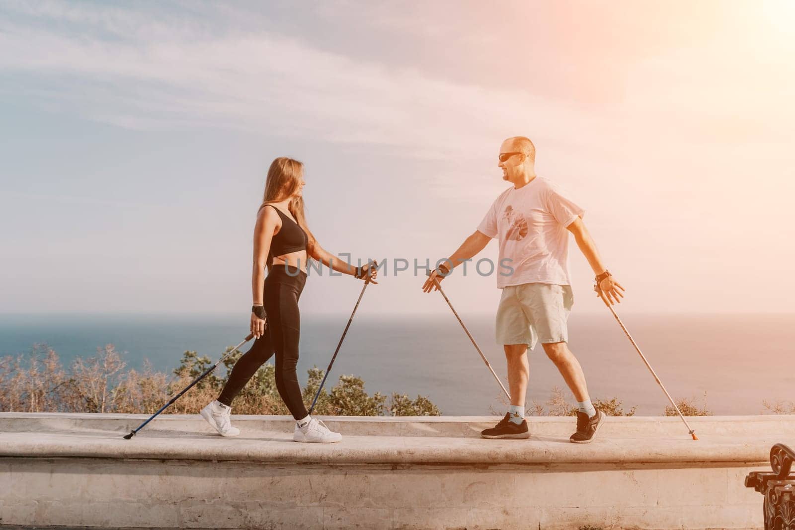 Happy Middle aged couple or friends practicing nordic walking in park near sea. Mature couple with trekking poles walking, practicing Nordic walking outdoors. Aging youthfully and sport concept by panophotograph