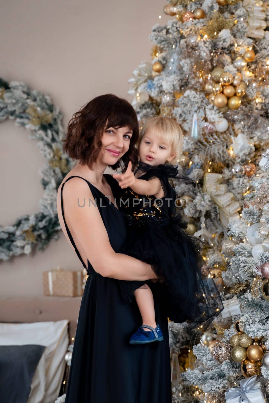 Mother daughter 2 years old Christmas tree. Both are dressed in black dresses, the mother holds the girl in her arms and both look at each other. The family celebrates Christmas