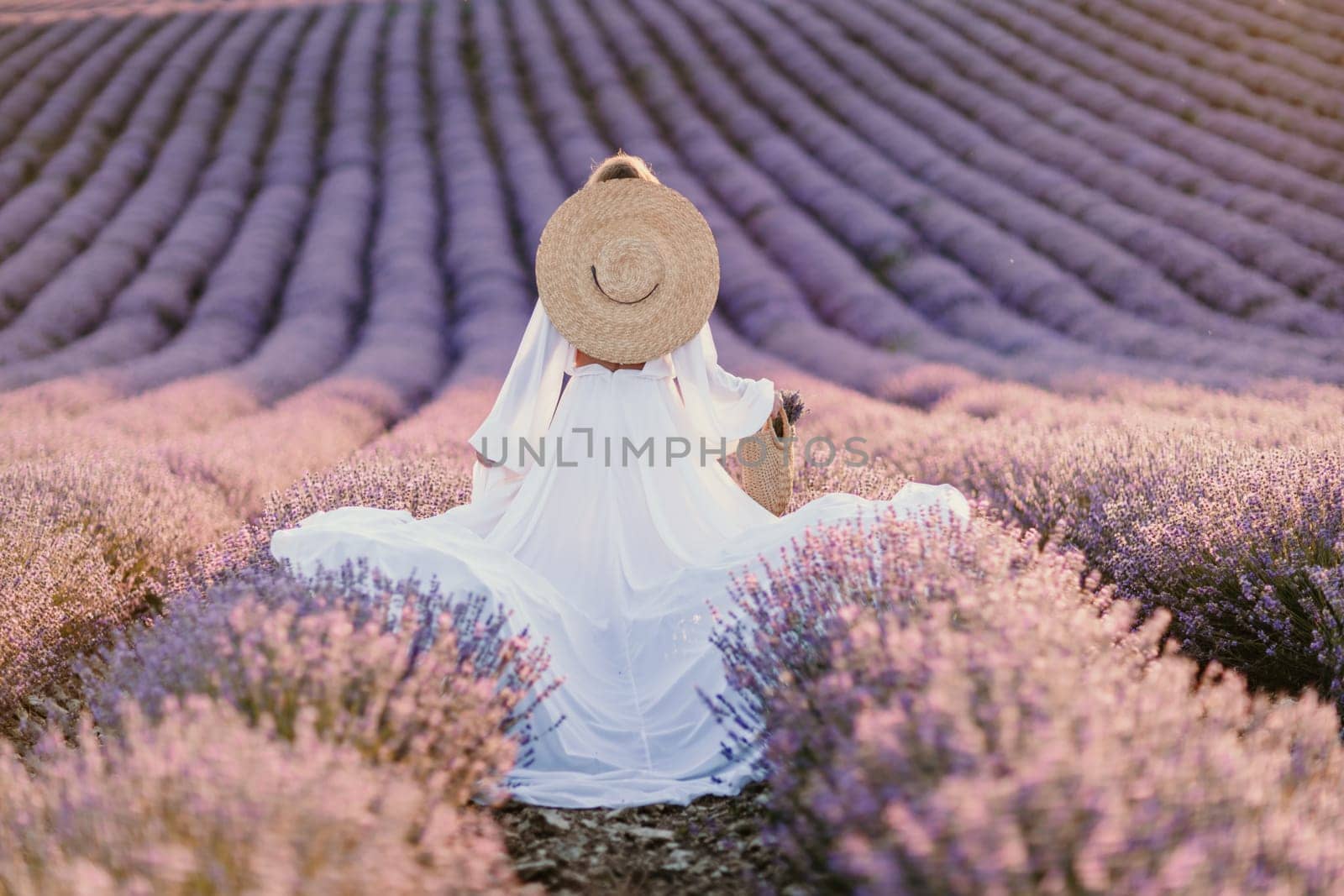 Happy woman in a white dress and straw hat strolling through a lavender field at sunrise, taking in the tranquil atmosphere. by Matiunina