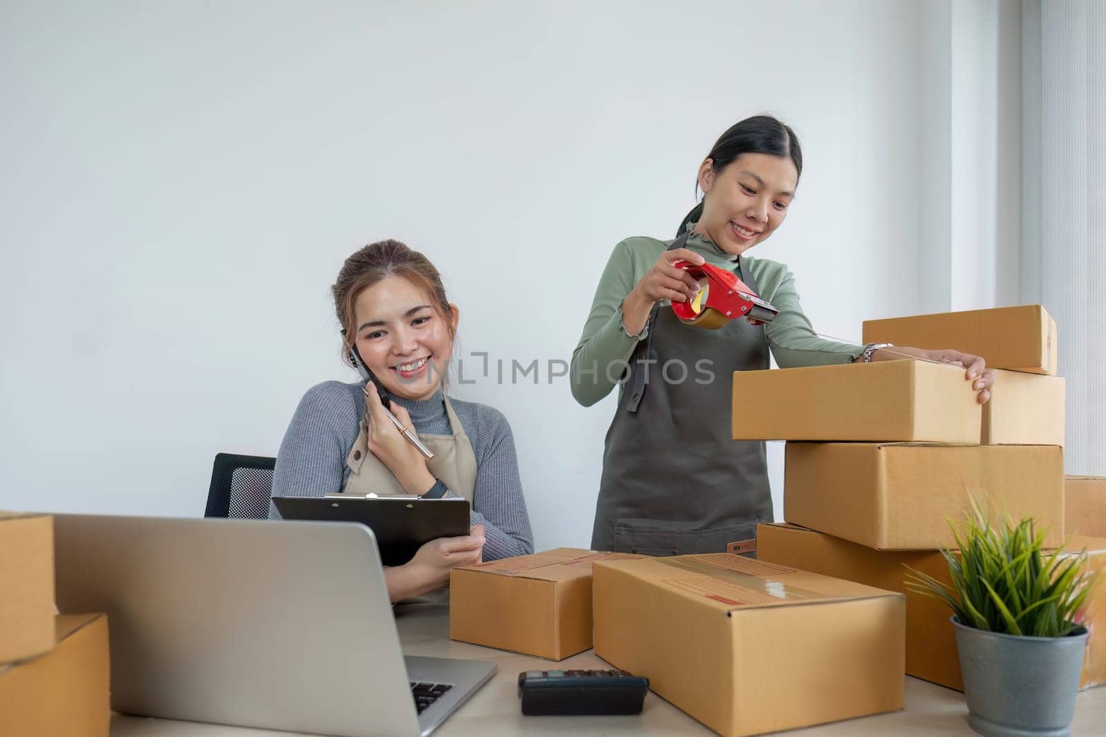 Two Asian entrepreneur women take order and check boxes of products according to customer orders in preparation for delivery. by wichayada