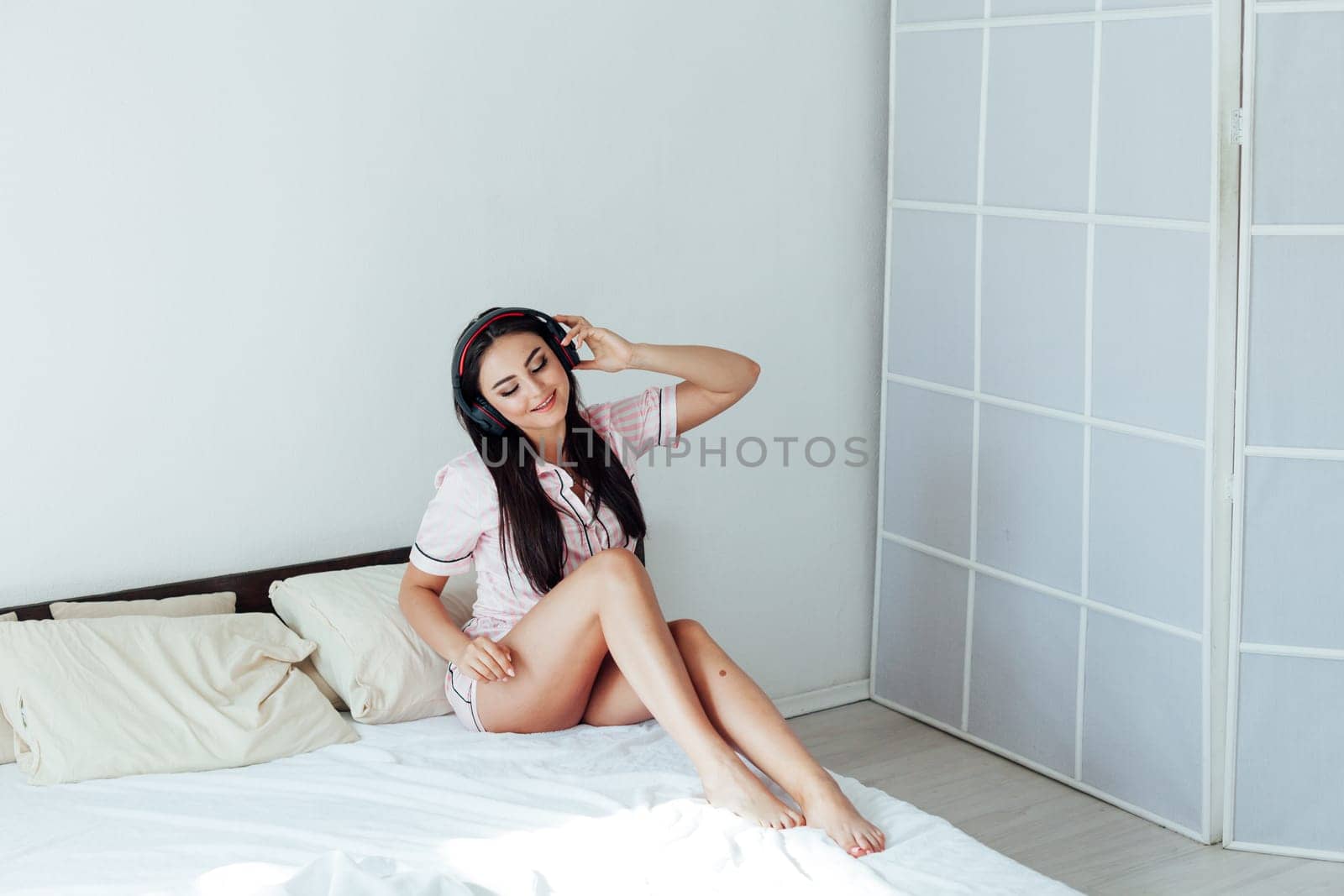 Woman in pajamas sitting on bed in the morning and listening to music with headphones by Simakov