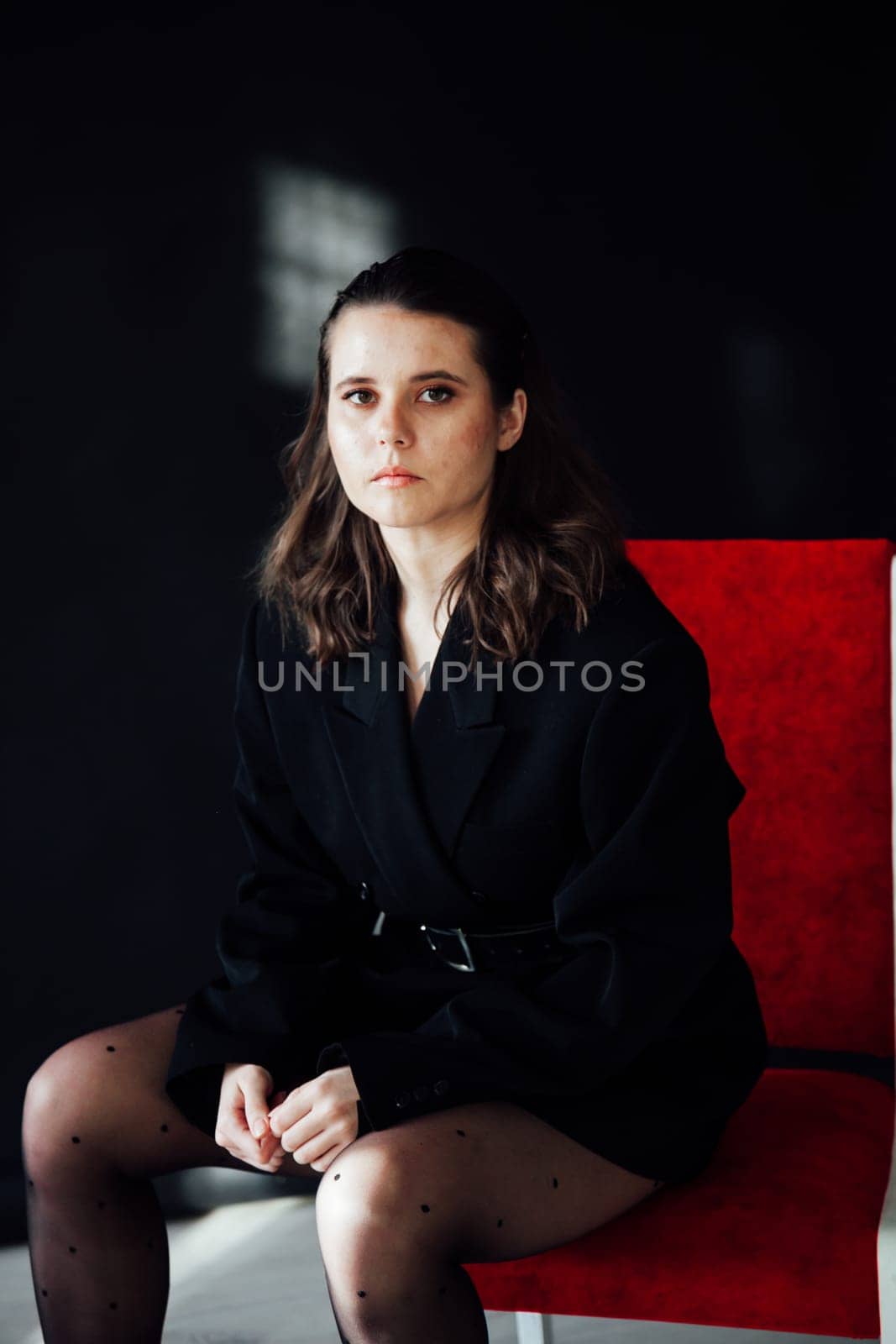 Fashionable brunette woman posing on black background in studio on red chair by Simakov