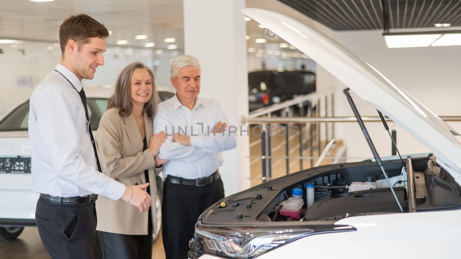 A salesman demonstrates a car with an open hood to an elderly Caucasian couple in a car dealership. by mrwed54
