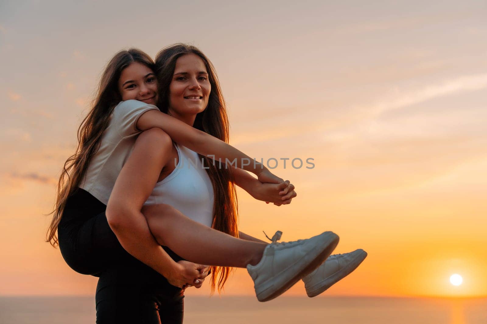 Mother carrying daughter on her back as they walk together, wearing a white shirt, and both are enjoying their time together, takes place during a sunset, adding a warm and serene atmosphere to the image. by Matiunina