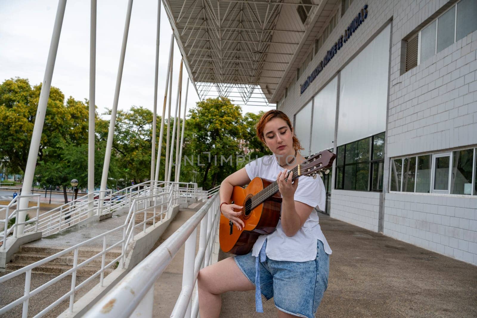 Side view of young female musician playing song on acoustic guitar while standing near metal fence of building on street in public place