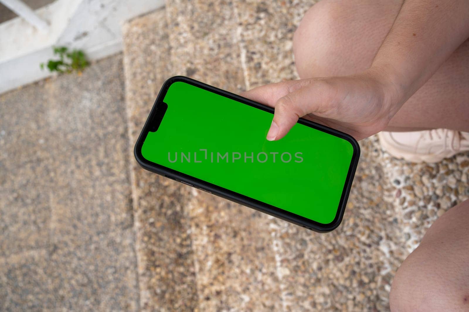 From above of anonymous young person in sneakers looking at chromakey screen of mobile phone and browsing messages while sitting on marble steps in daylight