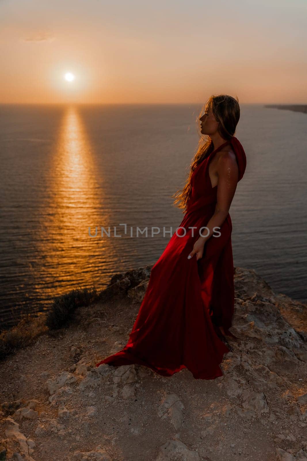 Woman sunset sea red dress, back view a happy beautiful sensual woman in a red long dress posing on a rock high above the sea on sunset