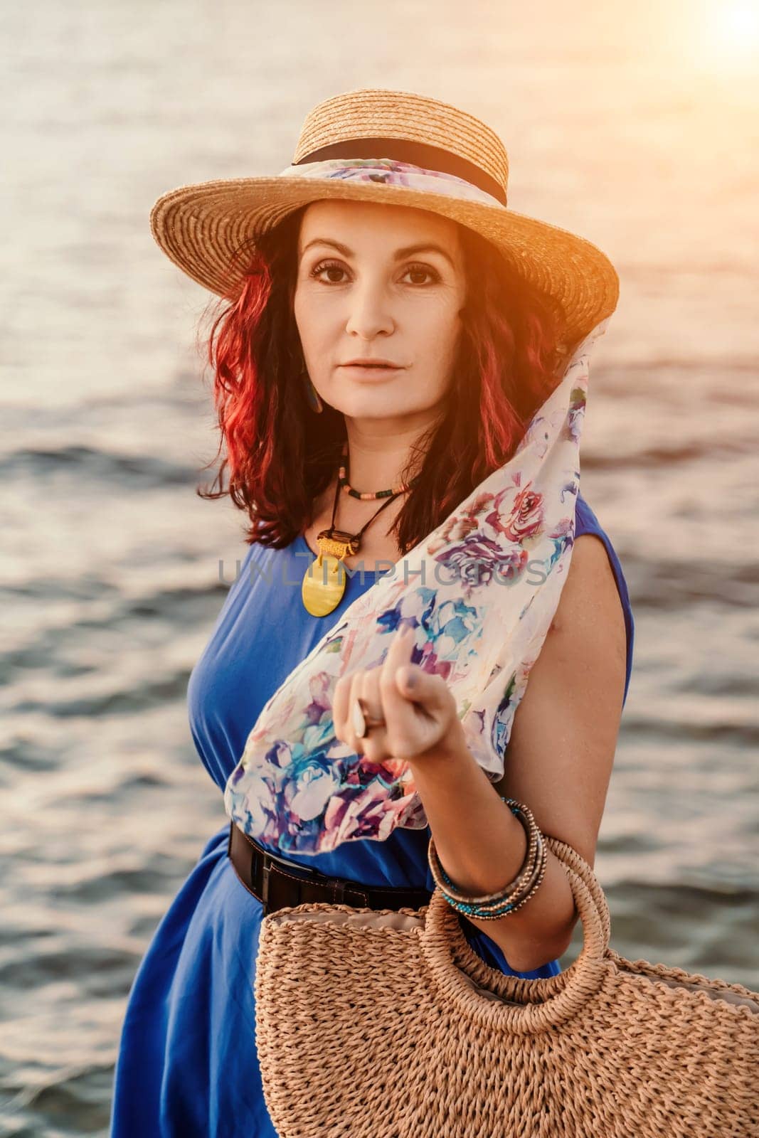 A woman in a dress, hat and with a straw bag is standing on the beach enjoying the sea. Happy summer holidays.