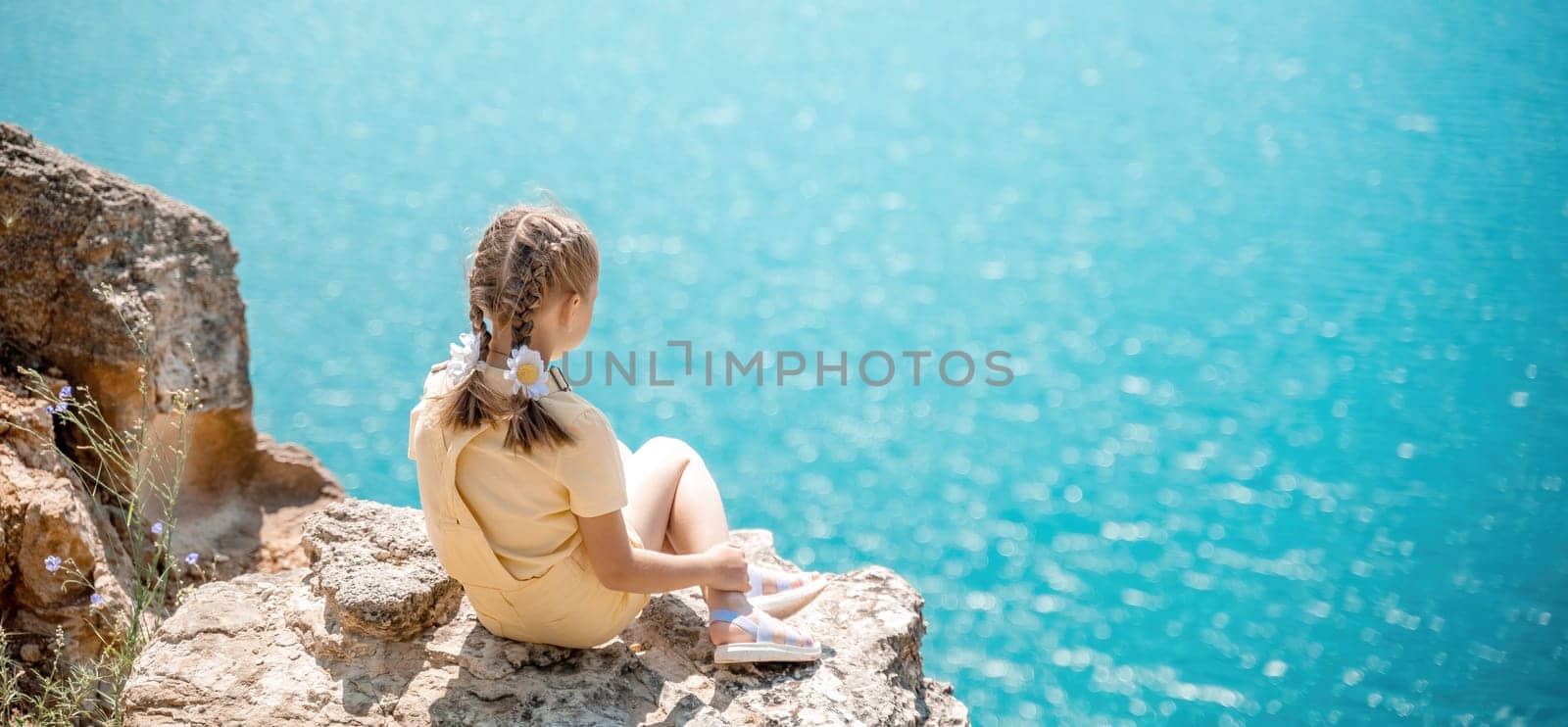 Happy girl perched atop a high rock above the sea, wearing a yellow jumpsuit and braided hair, signifying the concept of summer vacation at the beach. by Matiunina