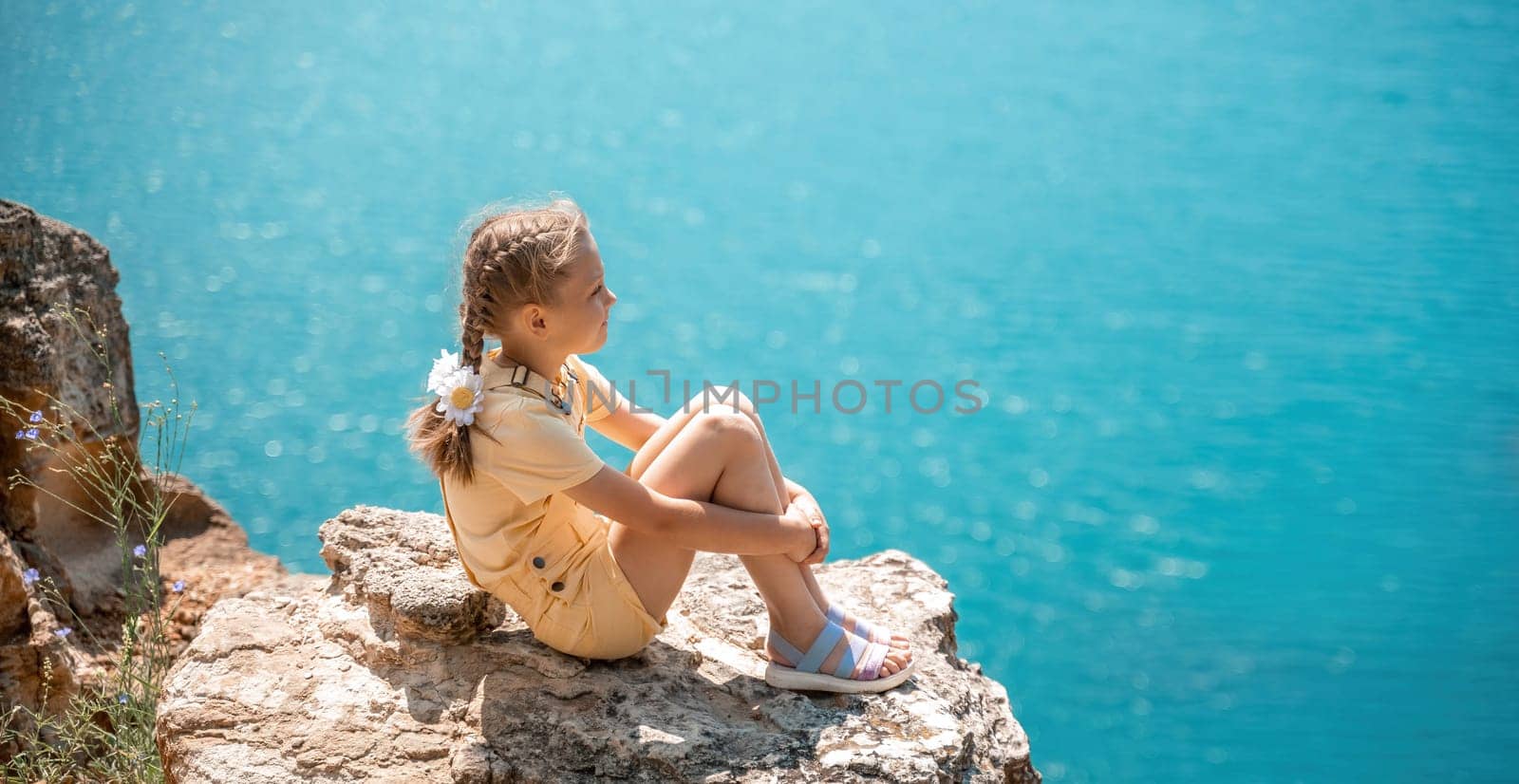 Happy girl perched atop a high rock above the sea, wearing a yellow jumpsuit and braided hair, signifying the concept of summer vacation at the beach. by Matiunina