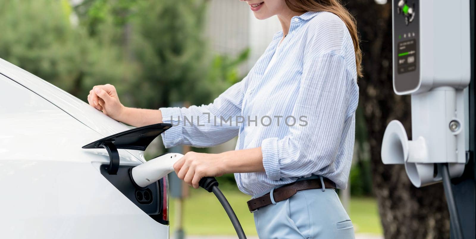 Young woman recharge EV electric vehicle's battery from EV charging station in outdoor green city park scenic. Eco friendly urban transport and commute with eco friendly EV car travel. Panorama Exalt