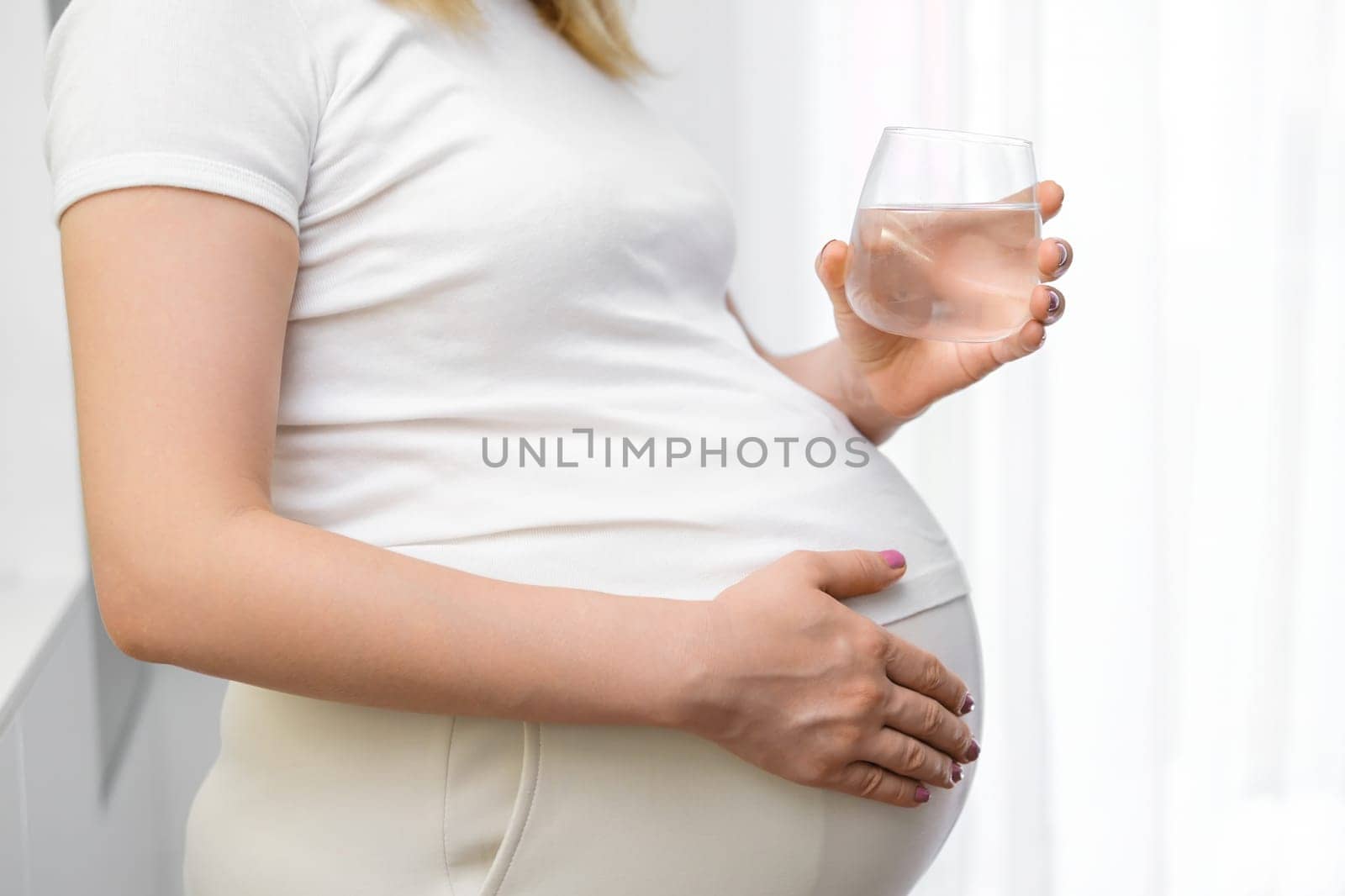 A pregnant woman holding glass of water in the kitchen. Hydration during pregnancy.