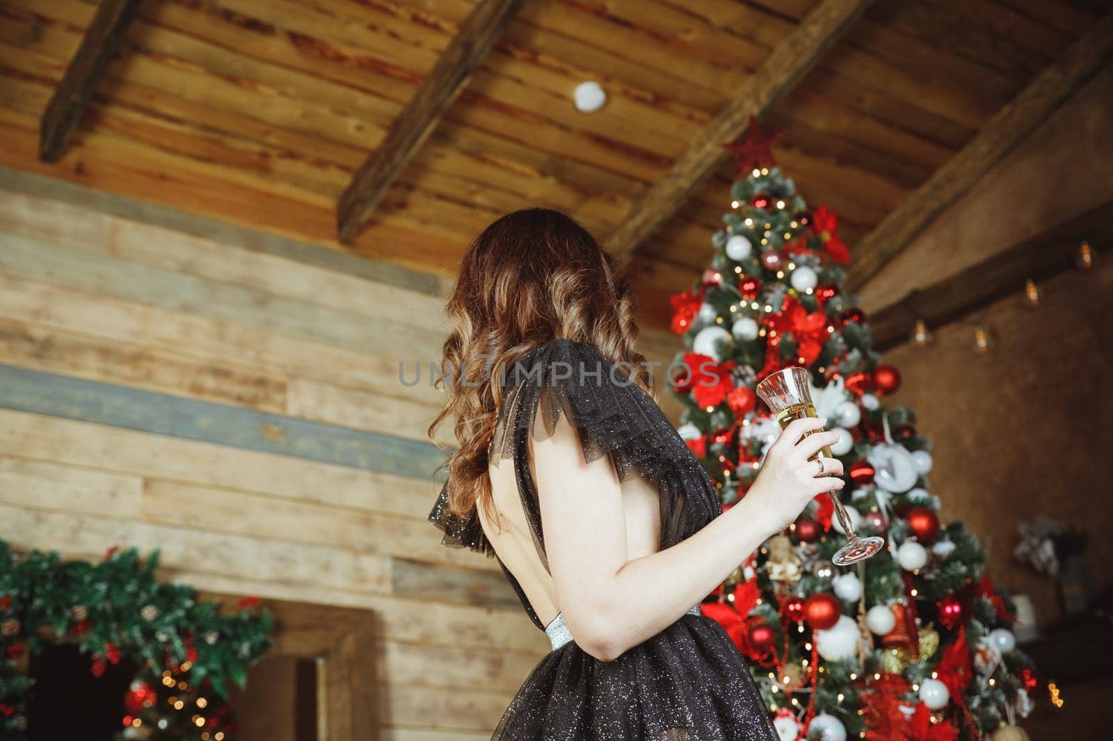 beautiful slender young brunette woman in black evening dress, stands next to decorated Christmas tree. Long slender legs.