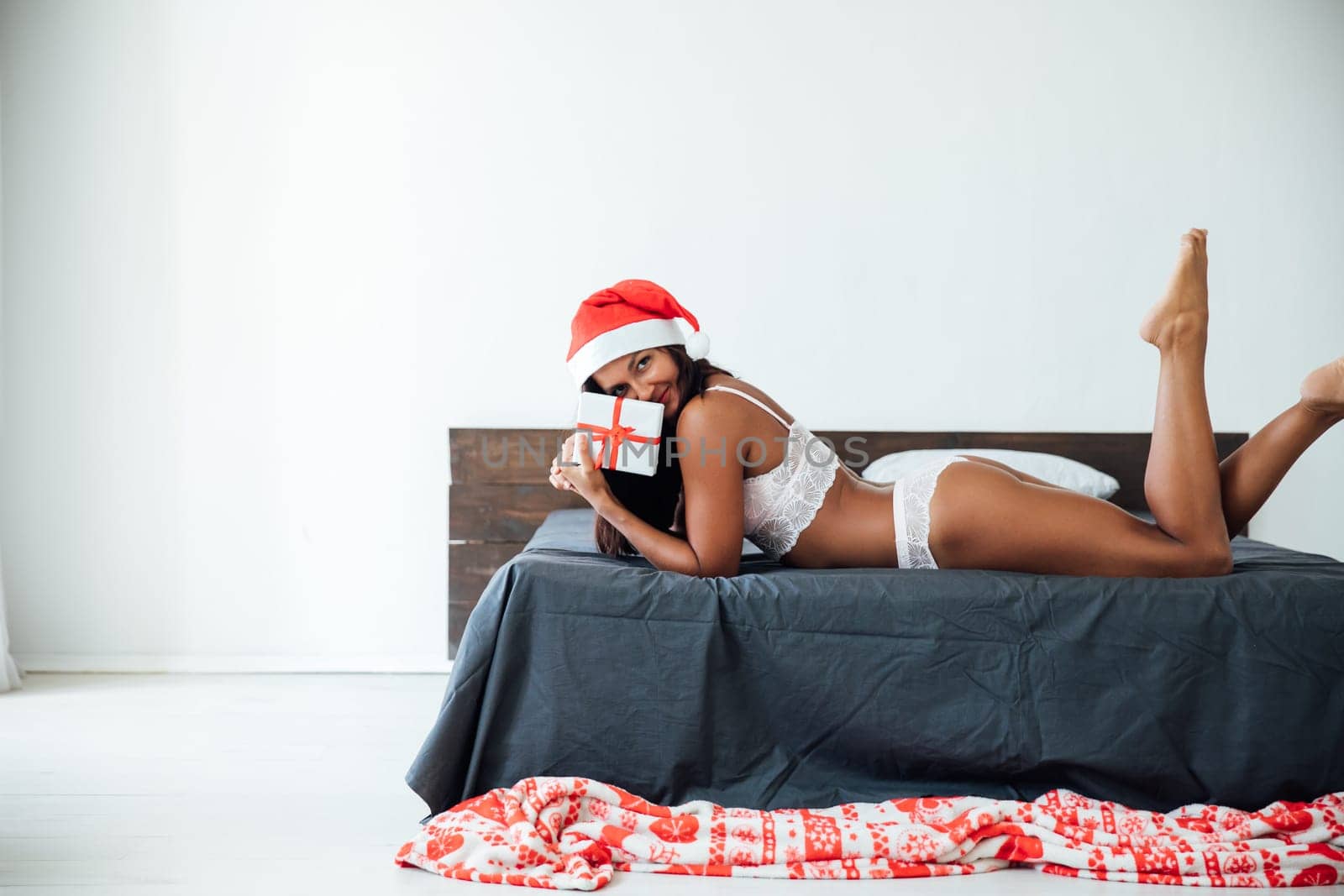 Holiday New Year Christmas Woman in Santa Claus hat with gift lying on the bed in the room by Simakov
